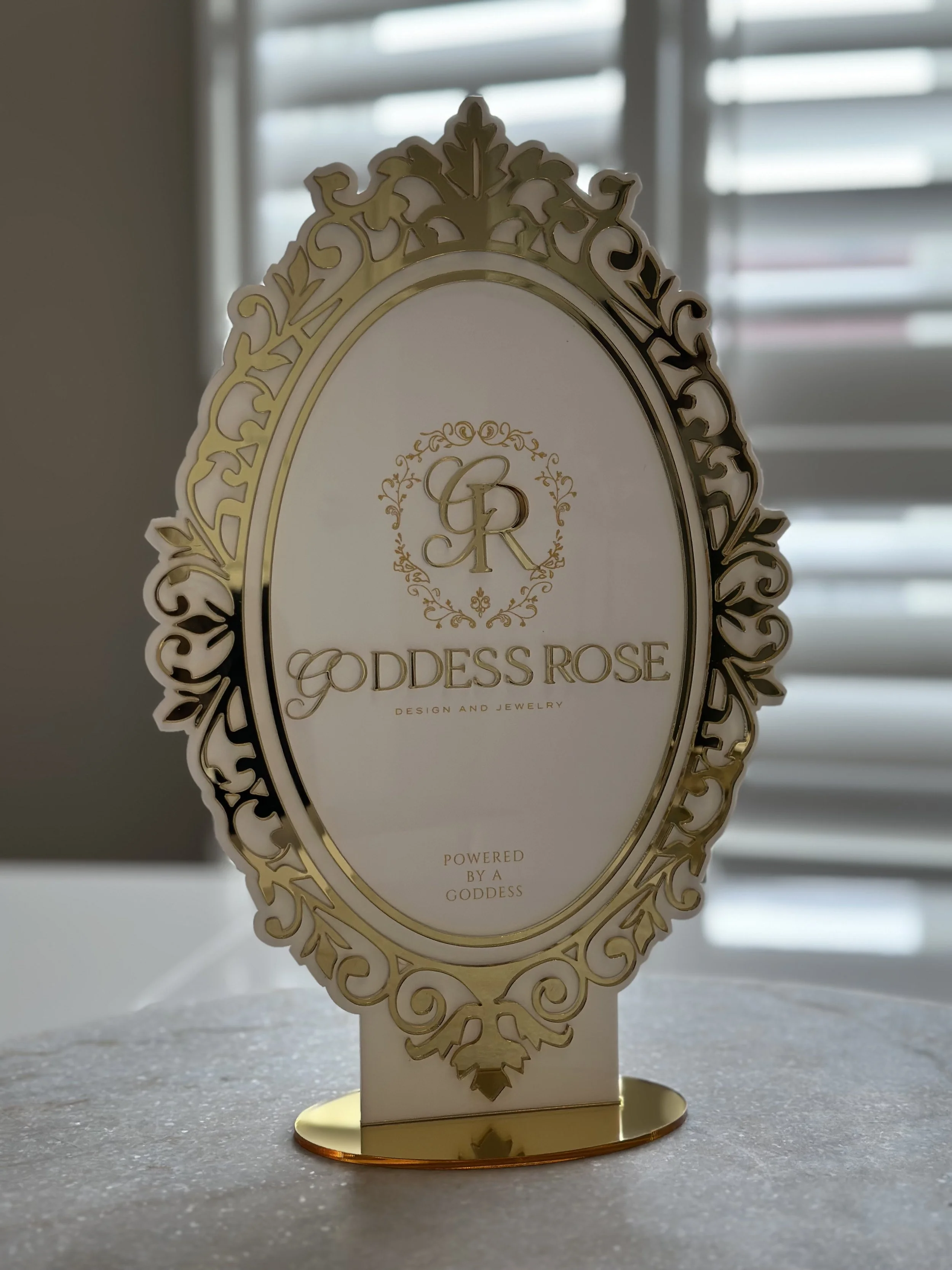 A decorative display with a white and gold ornate oval frame, featuring the words 'Goddess Rose' in gold lettering, along with a monogram 'GR' and the phrase 'Powered by a Goddess' at the bottom.