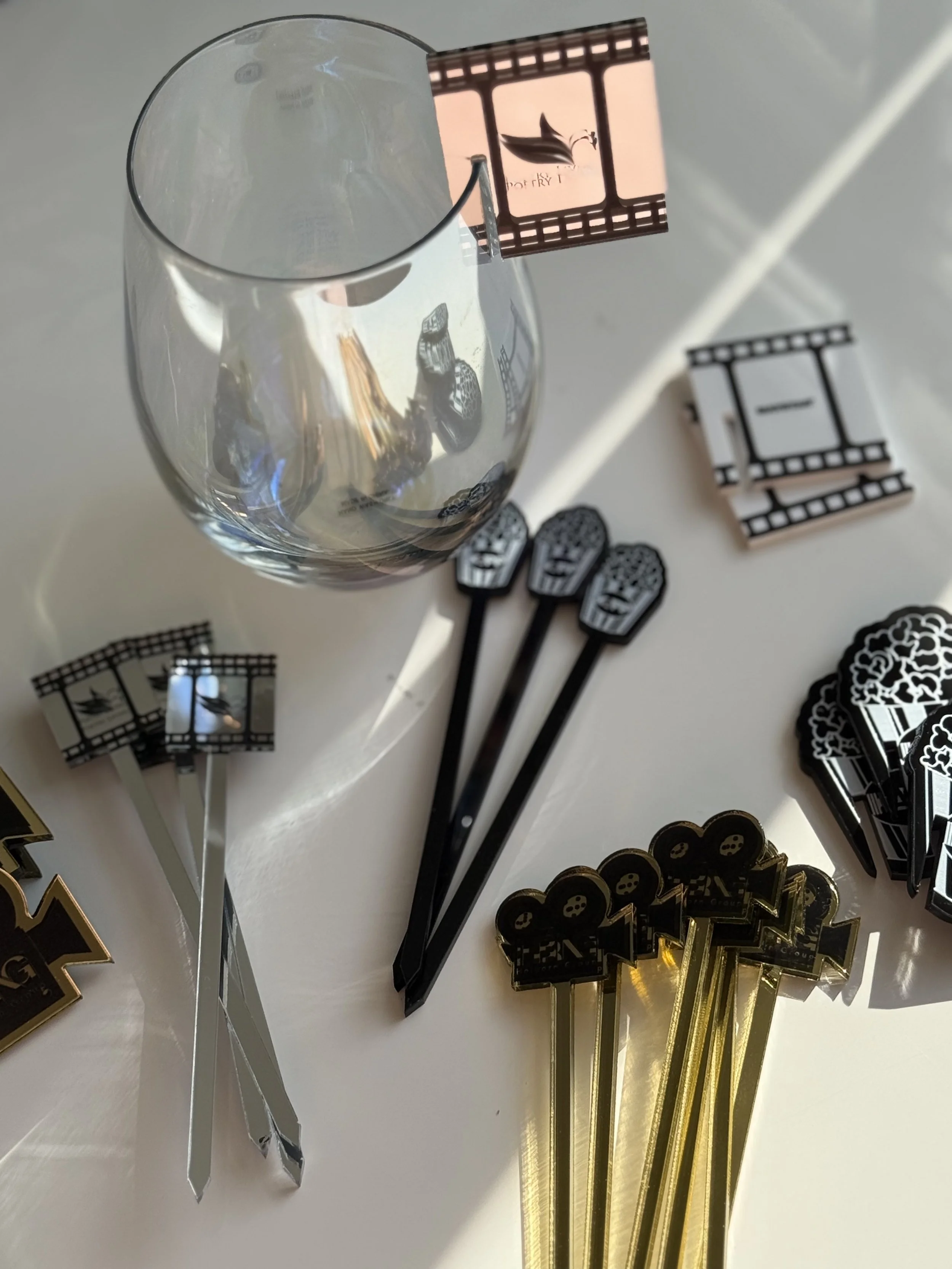 Plastic black and gold-themed drink stirrers, some with camera designs, some with film reels, and one with a bird design, scattered on a white surface next to an empty wine glass.