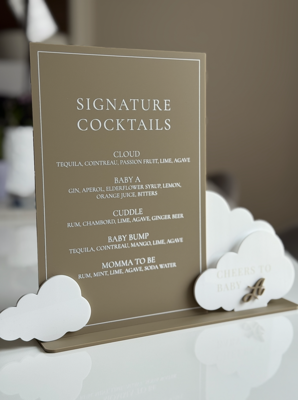 A menu card listing signature cocktails, with cloud-shaped decorations on a white table, at a celebratory event.