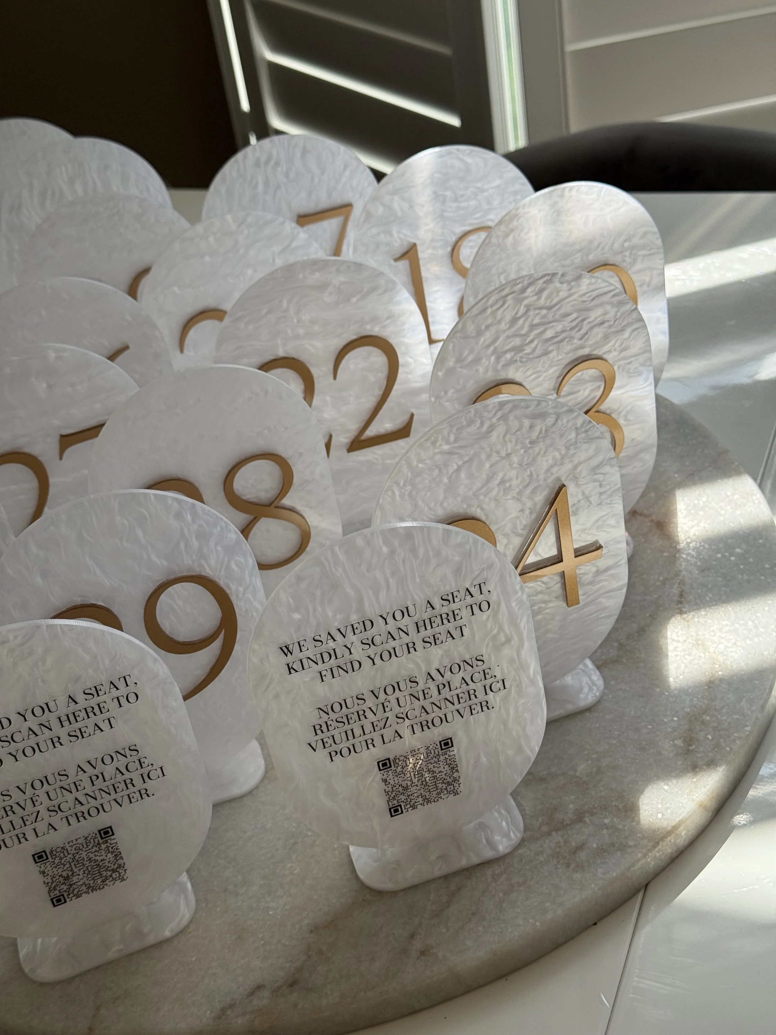 White table number signs with gold numbers and an instructions card with a QR code on a marble surface.