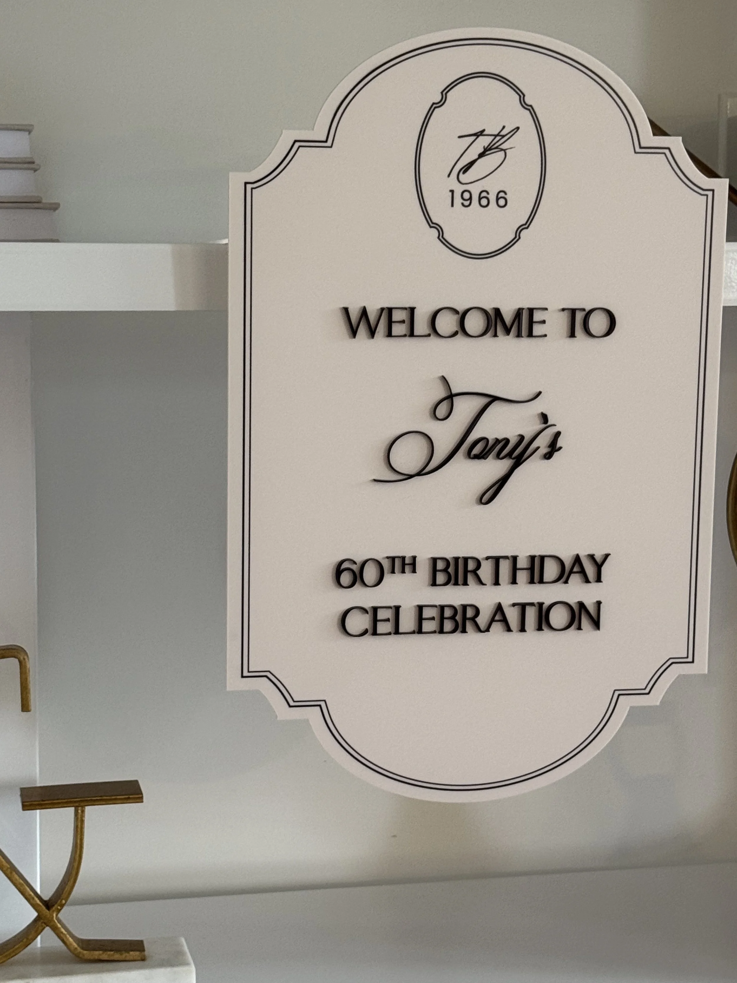 A decorative white sign with black border, featuring a scripted '76' and '1966' inside an oval at the top, and text that reads 'WELCOME TO Tony's 60TH BIRTHDAY CELEBRATION' in elegant fonts.