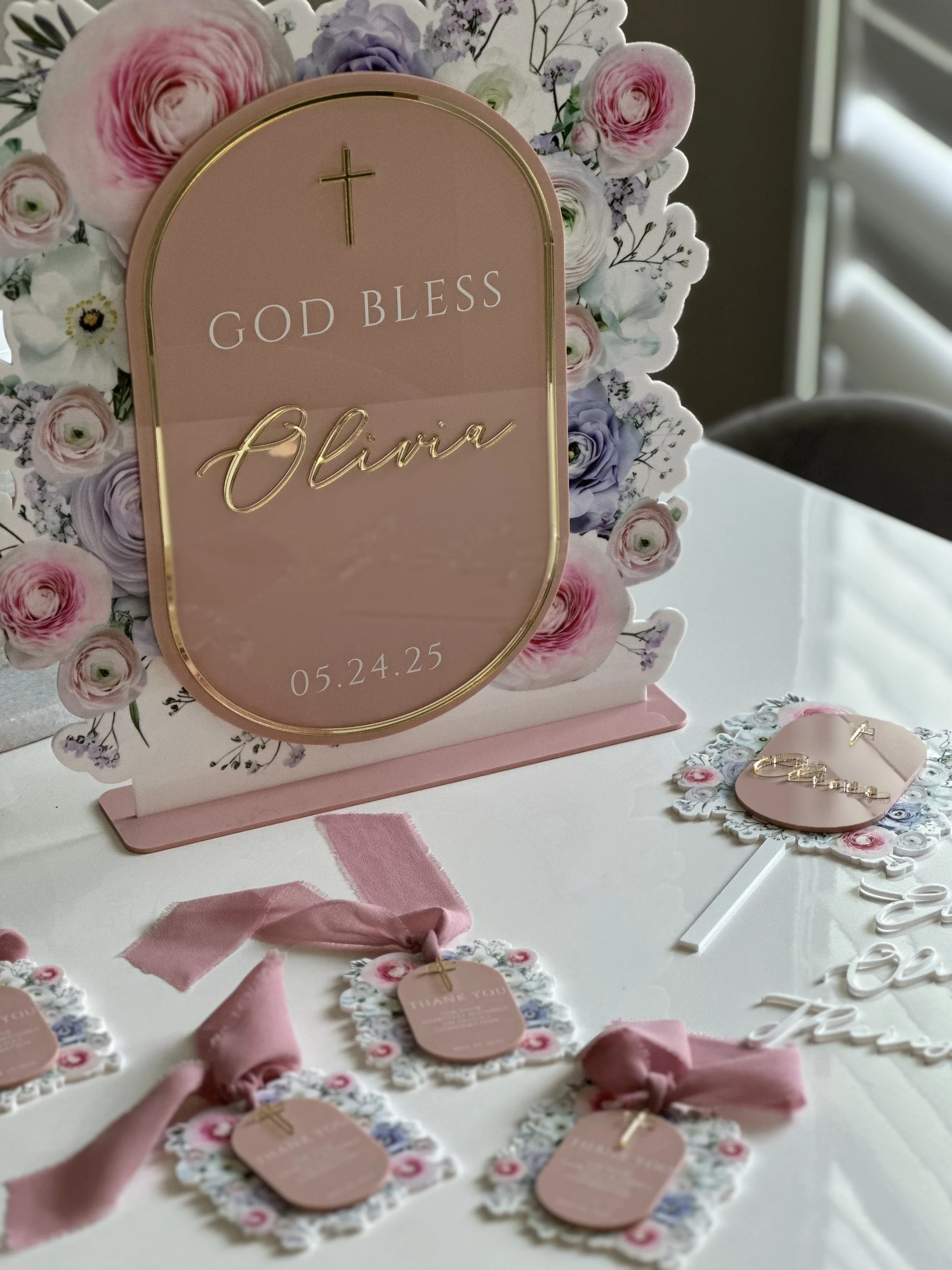 A pink and floral-themed wedding or event decoration with a sign that says "God Bless Olivia" and the date 05.24.25. The decoration includes a gold cross at the top and surrounding floral elements in pastel colors. There are also small tags on pink r