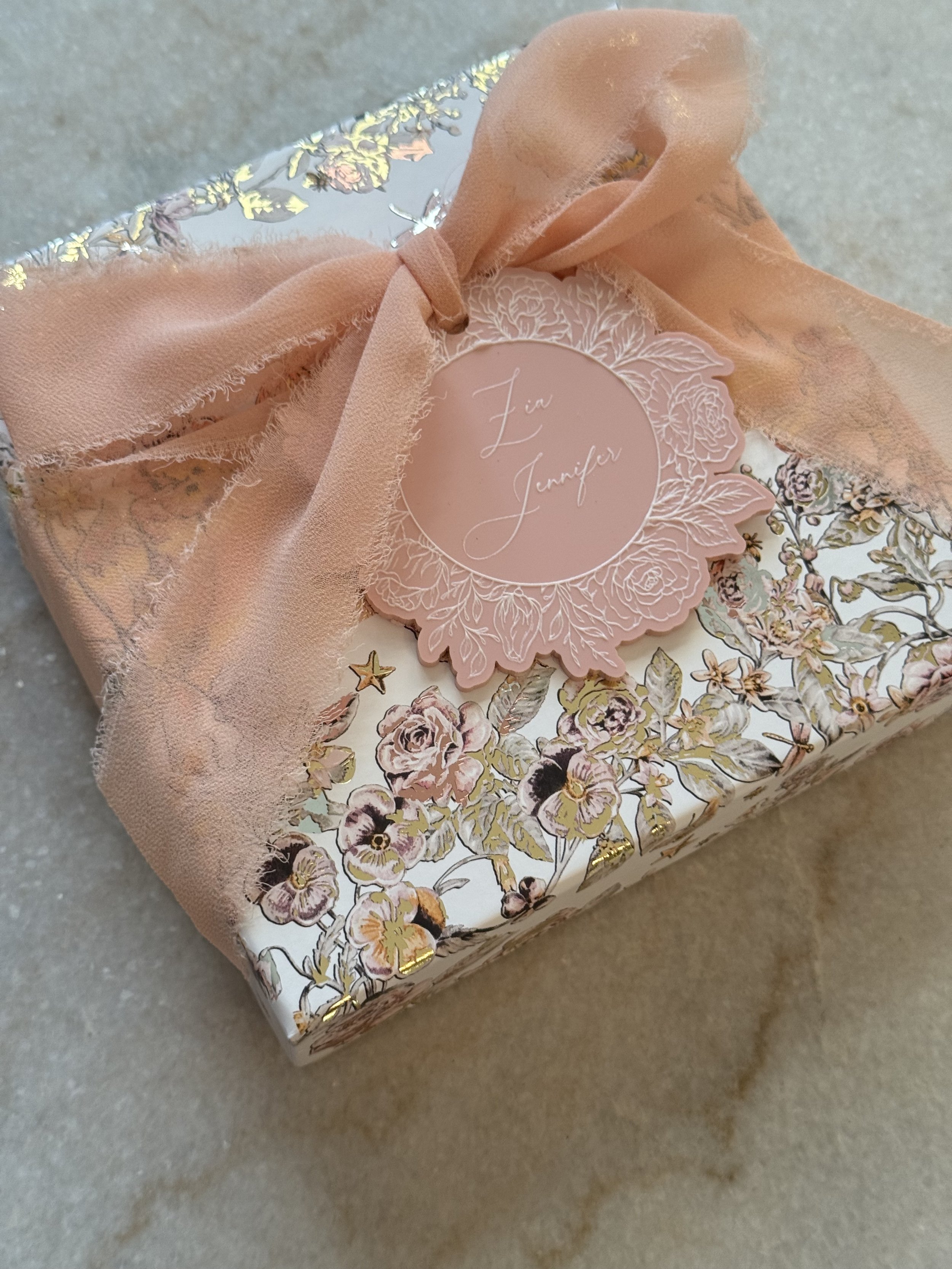 Decorative gift box with floral pattern, pink ribbon bow, and a round pink tag with the names 'Liz & Jennifer' on it.