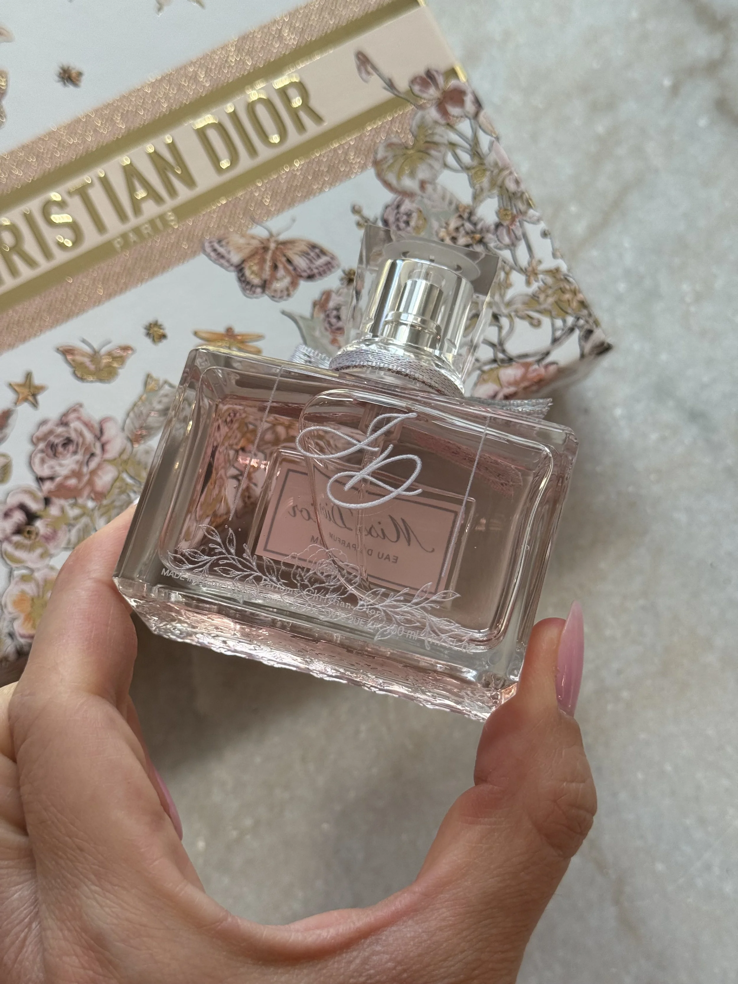 A hand holding a pink perfume bottle with floral and cursive design, with a Christian Dior gift box decorated with butterflies, flowers, and stars in the background.