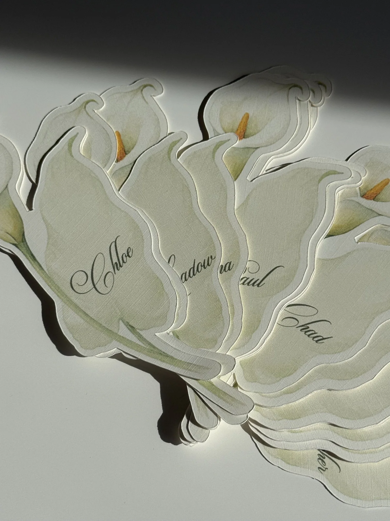 A collection of white paper cutouts shaped like calla lilies with orange stamens, layered to create a floral design with partially visible cursive handwriting.