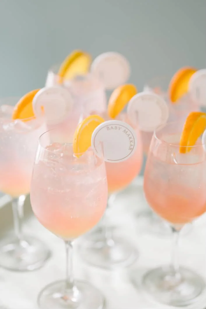 Multiple pink cocktails in stemmed glasses, garnished with lemon wedges and round white labels, on a white surface against a light background.