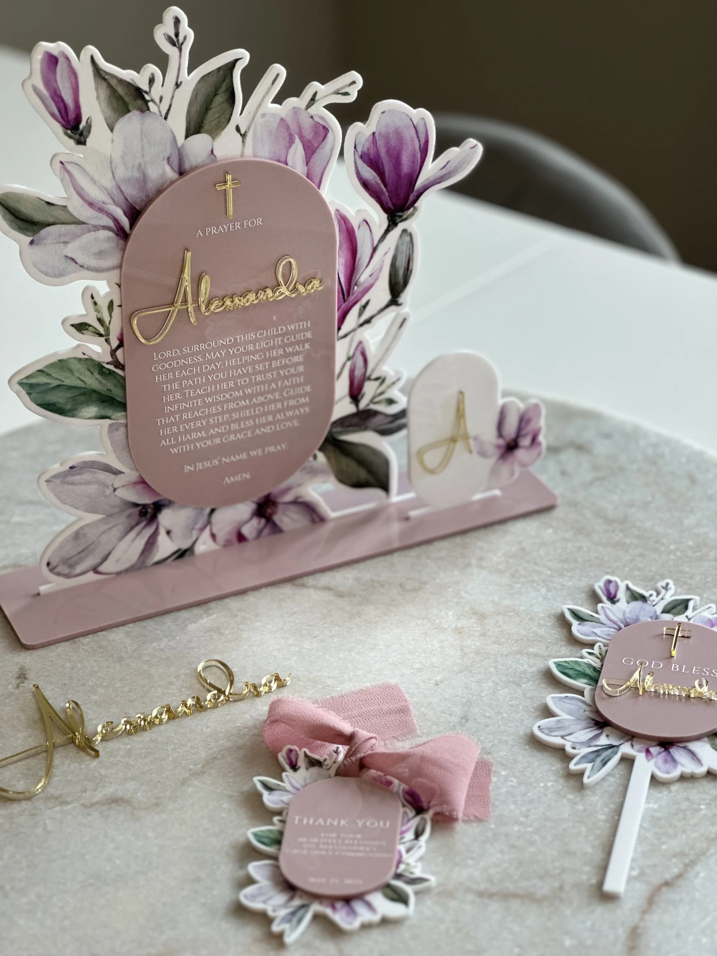 Decorative Christian-themed table setup with a floral photo stand featuring prayer and blessing messages, pink and purple flower designs, a pink thank you tag with a pink bow, and a gold 'Jasmine' nameplate.