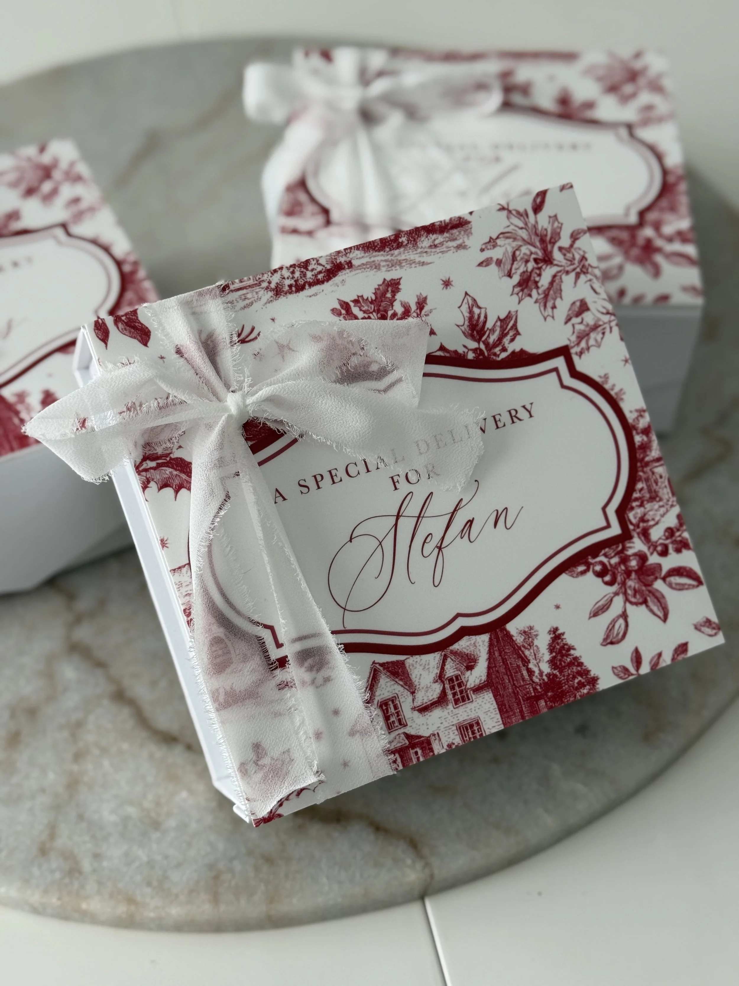 Close-up of a holiday gift box with white ribbon decoration, featuring red and white festive design, and a label reading 'A special delivery for Stefan'.