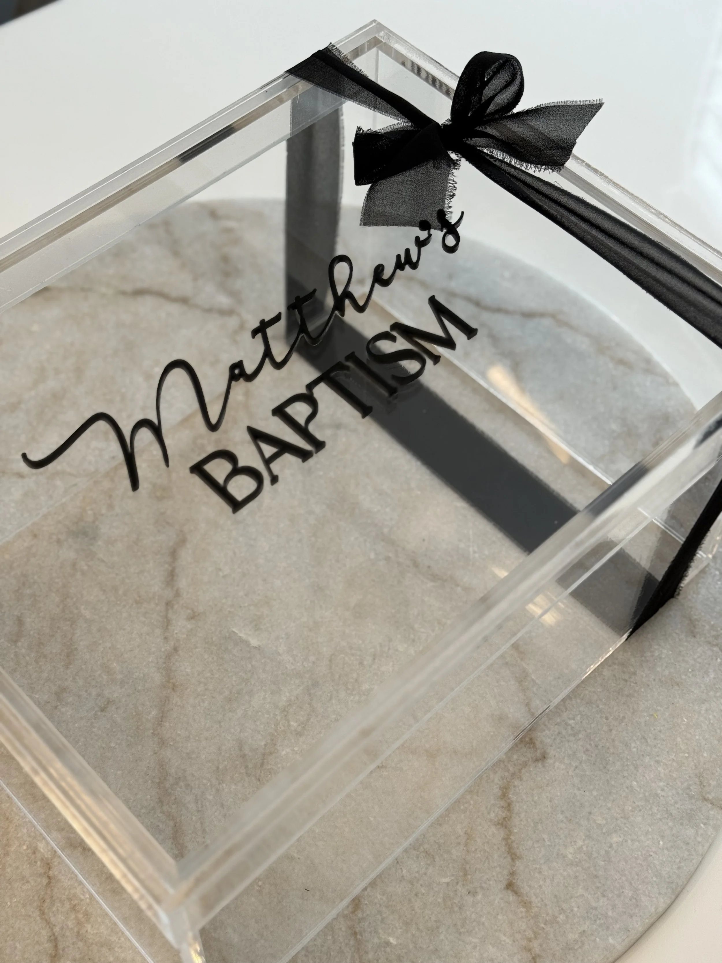 A clear acrylic box with black lettering that reads 'motherew B APSIM' and a black ribbon bow on top, placed on a light-colored stone surface.