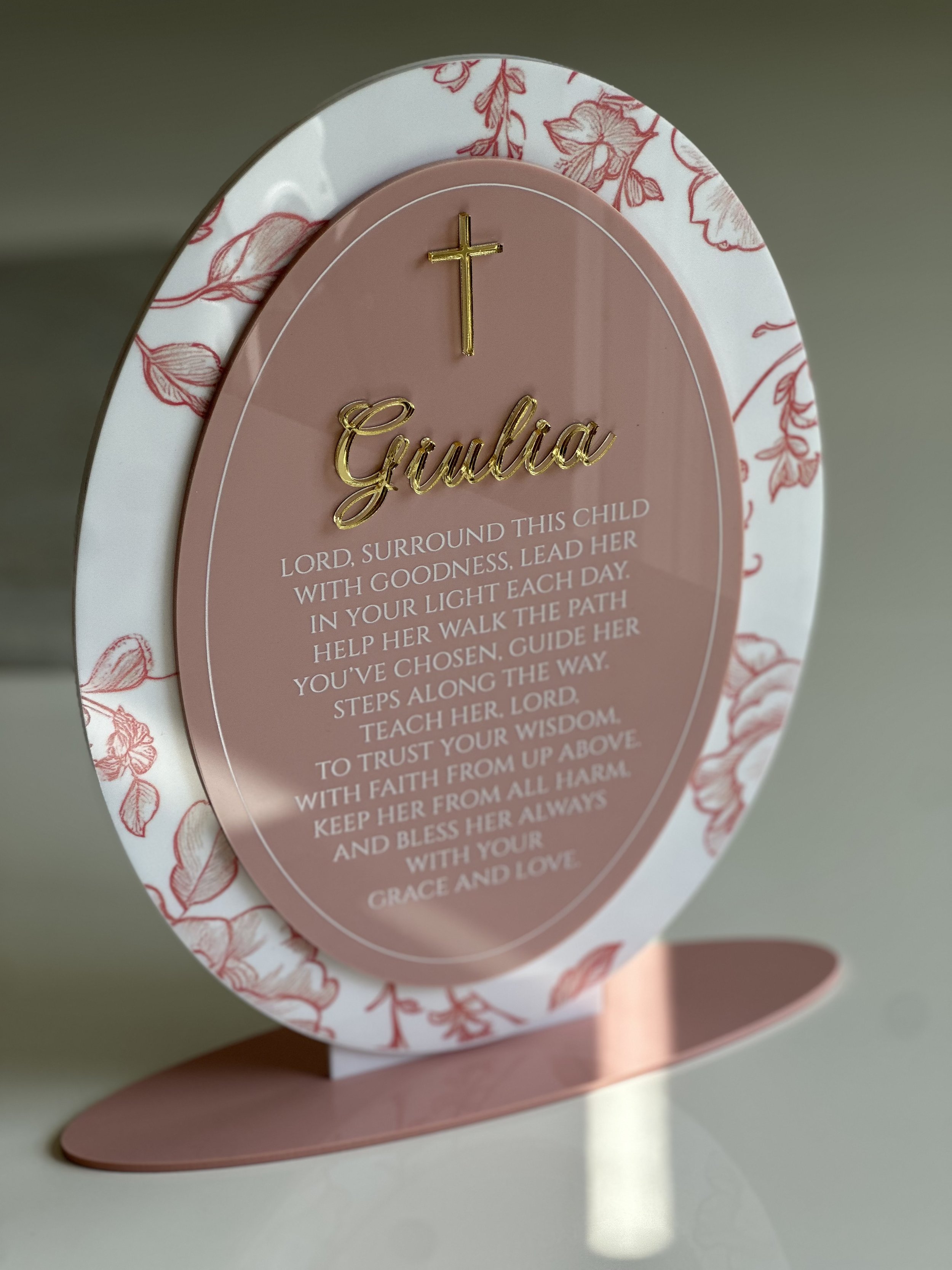 Decorative prayer plaque with a pink background, a gold cross, the name 'Gudia' in gold, and a prayer text, surrounded by a matching floral border, placed on a pink stand.