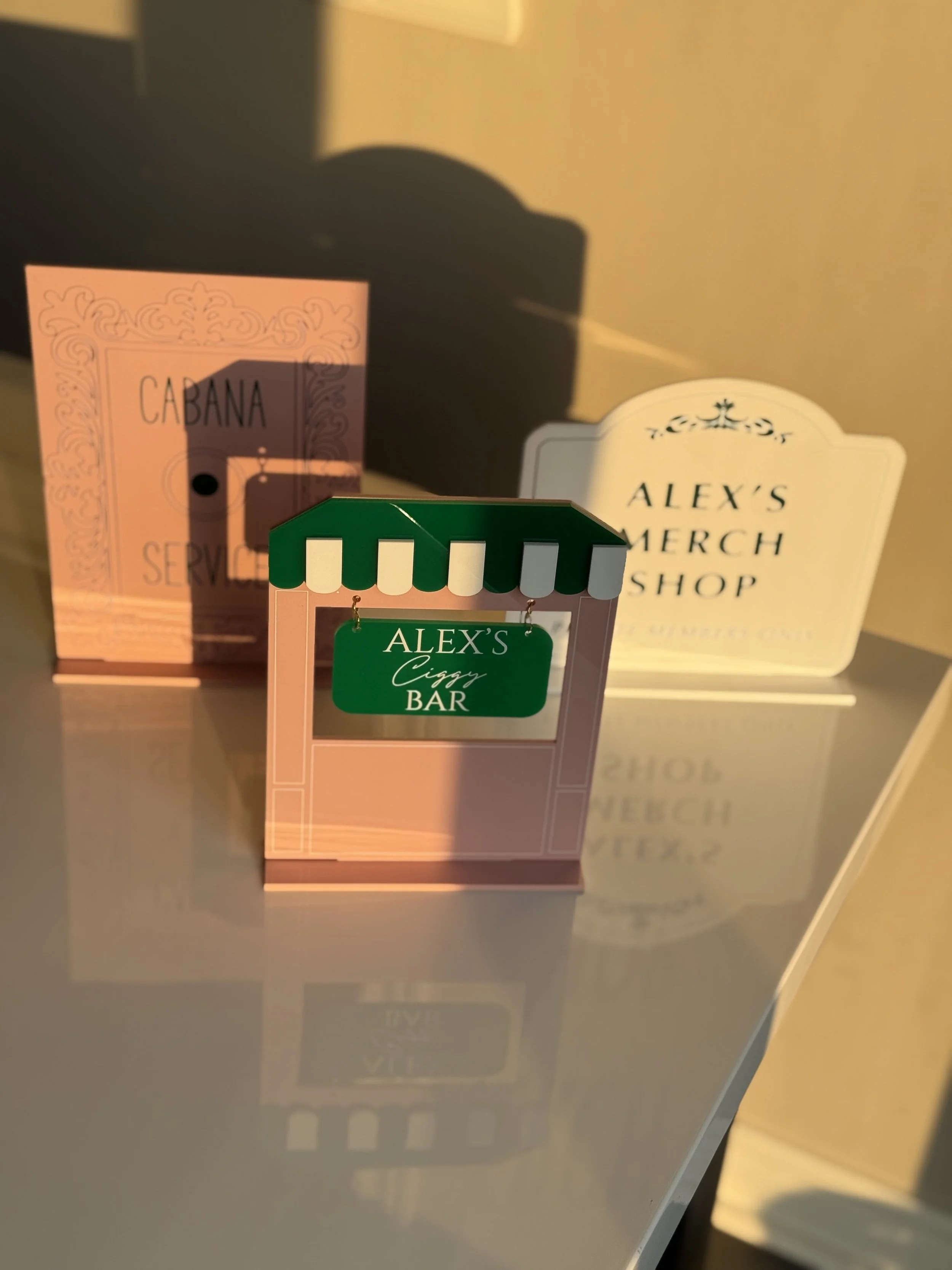 Miniature signboard with green and white canopy that reads 'ALEX'S CIGGY BAR' in front of two other signs, one pink with black text and one white with black and gold text, on a reflective surface.