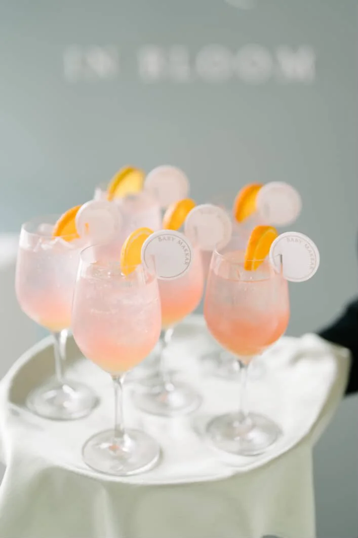 Six pink cocktails in coupe glasses garnished with orange slices and circular toppers, on a white tray with a blurred background.