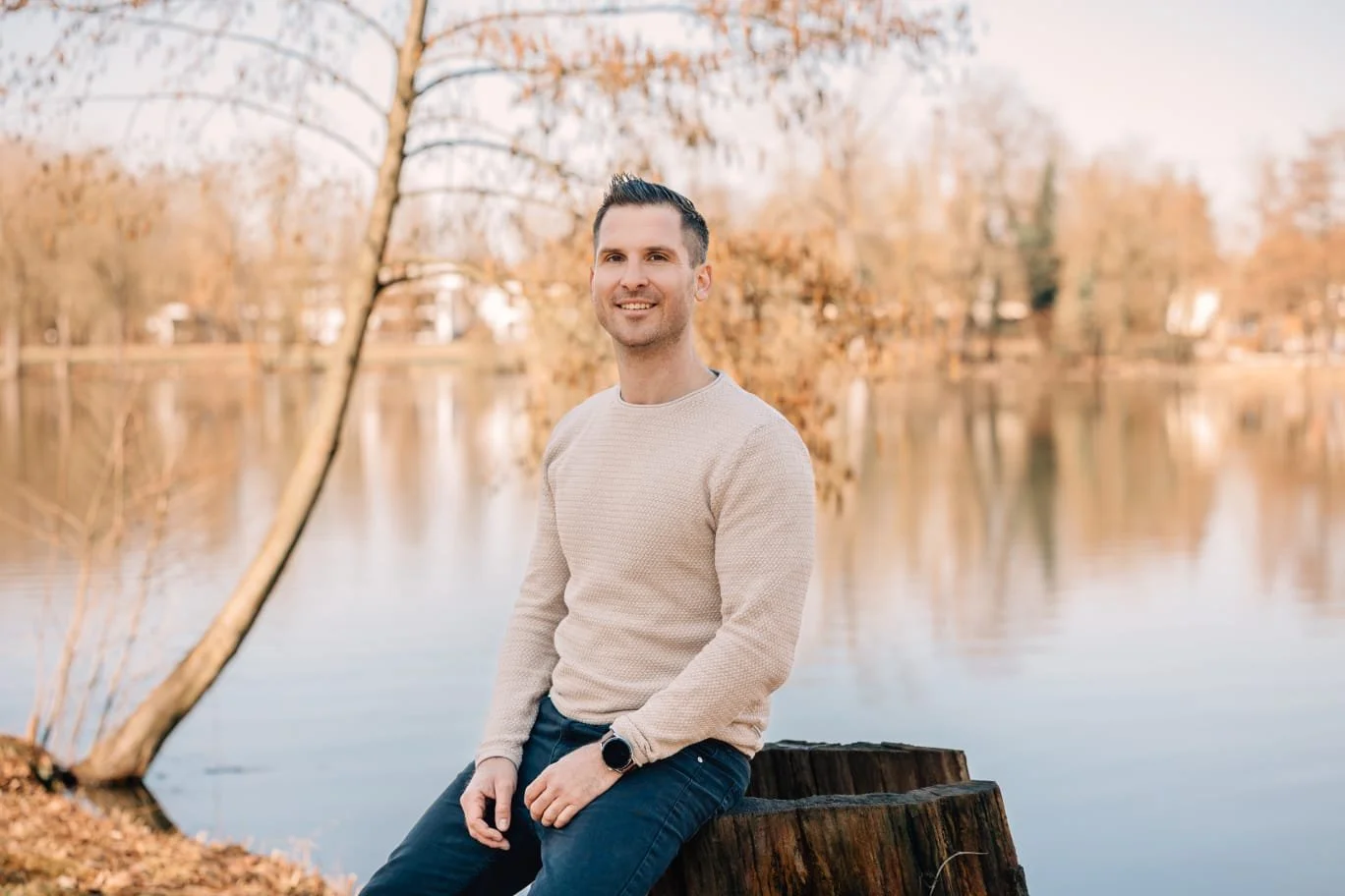 Junge Mann sitzt auf einem Baumstumpf am Fluss, umgeben von Herbstbäumen mit braunen Blättern, im Freien bei Sonnenuntergang.