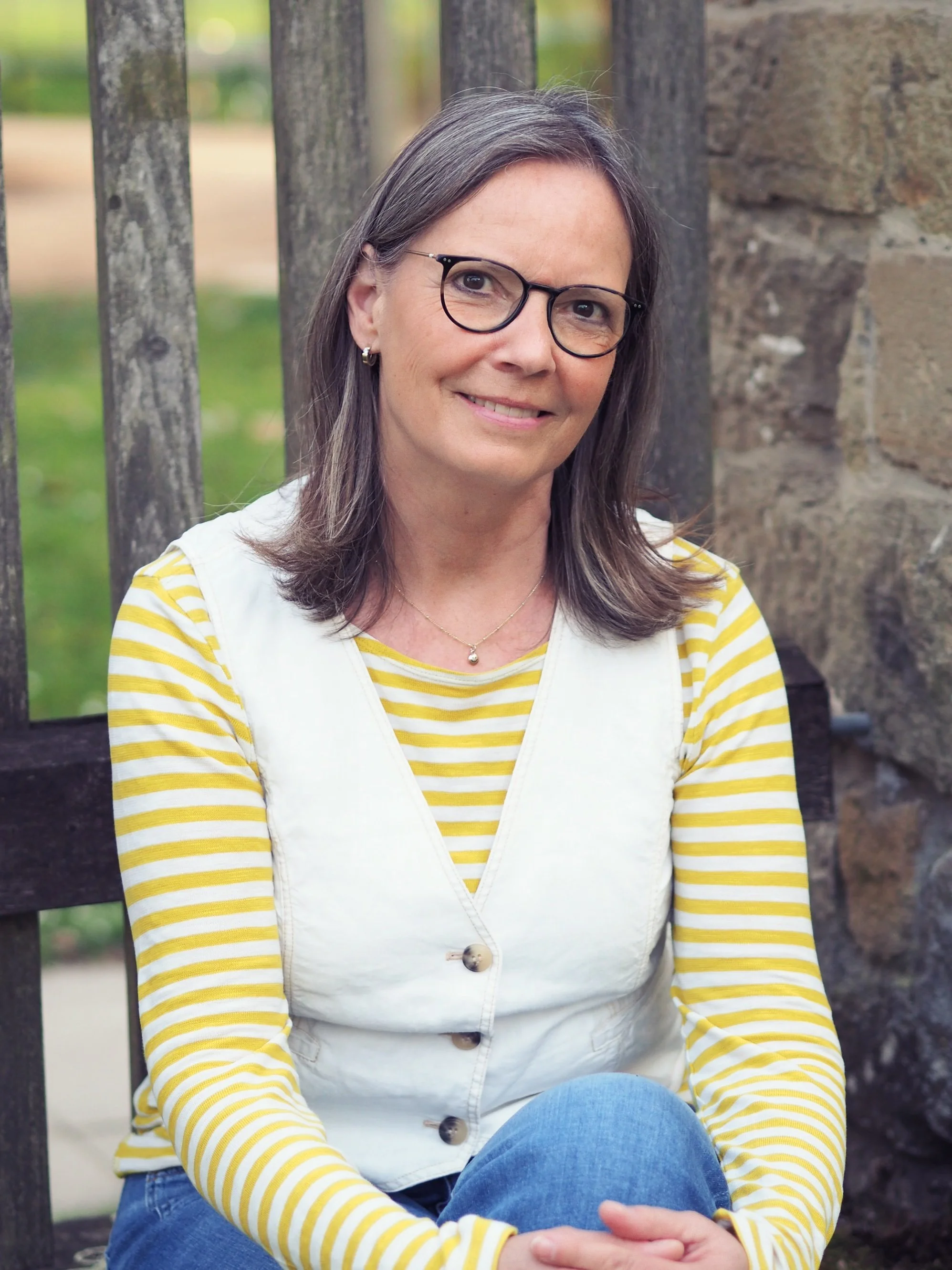 Eine lächelnde Frau mit Brille sitzt im Freien vor einer Holzzaun und einer Steinmauer, trägt ein gelb-weiß gestreiftes Oberteil und eine weiße Weste.