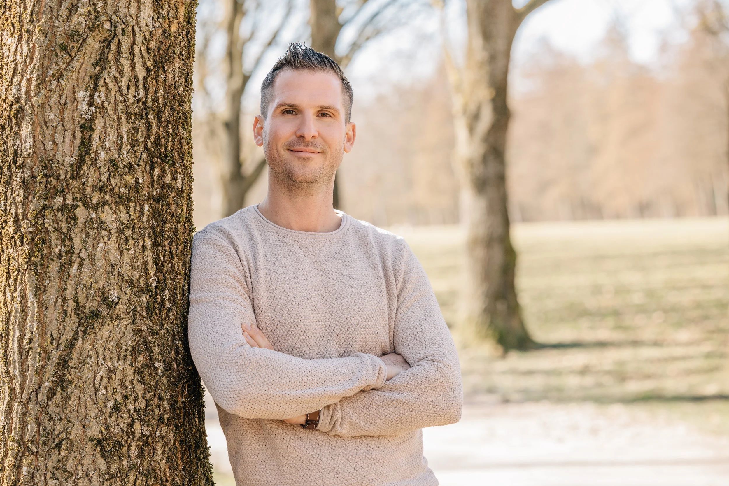 Ein Mann steht im Freien neben einem Baum, trägt einen hellen Pullover, lächelt leicht und hat die Arme vor der Brust.
