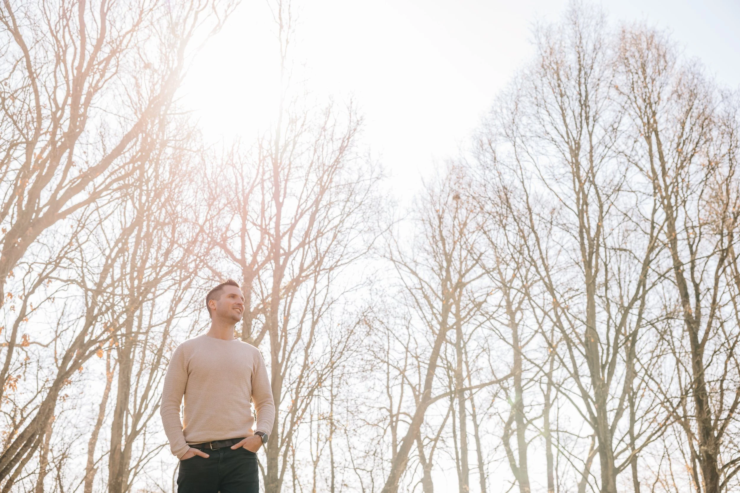 Mensch mit ordentlich geschnittenem Haar in einem beige Pullover und dunklen Hosen steht in einem Wald mit kahl gewordenen Bäumen, die Zweige sind sichtbar, und der Himmel ist hell mit starker Sonneneinstrahlung.