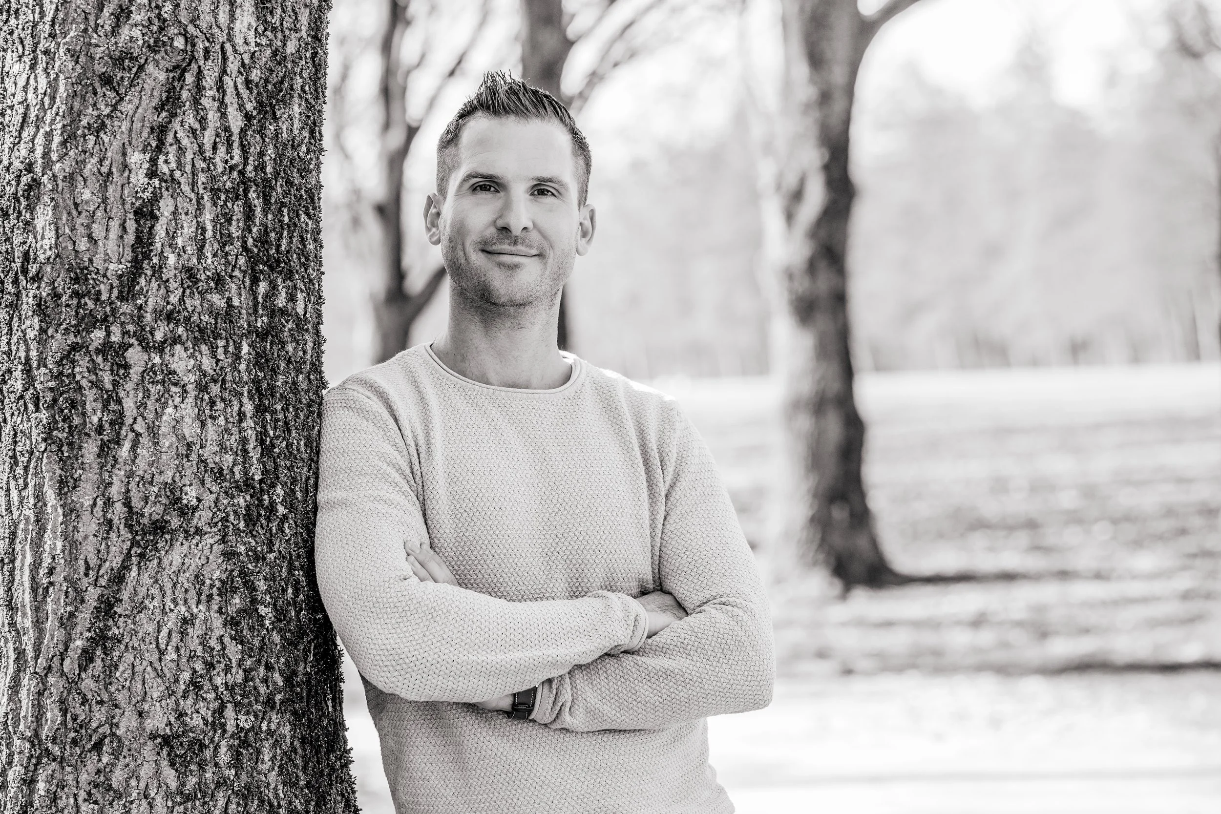 Ein Mann steht mit verschränkten Armen neben einem Baum im Park, lächelt leicht und blickt in die Kamera, im Hintergrund sind weitere Bäume zu sehen, alles in Schwarz-Weiß.