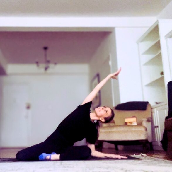 A woman practicing yoga on a carpeted floor in a living room, leaning to one side with one arm extended upward and the other resting on the floor.
