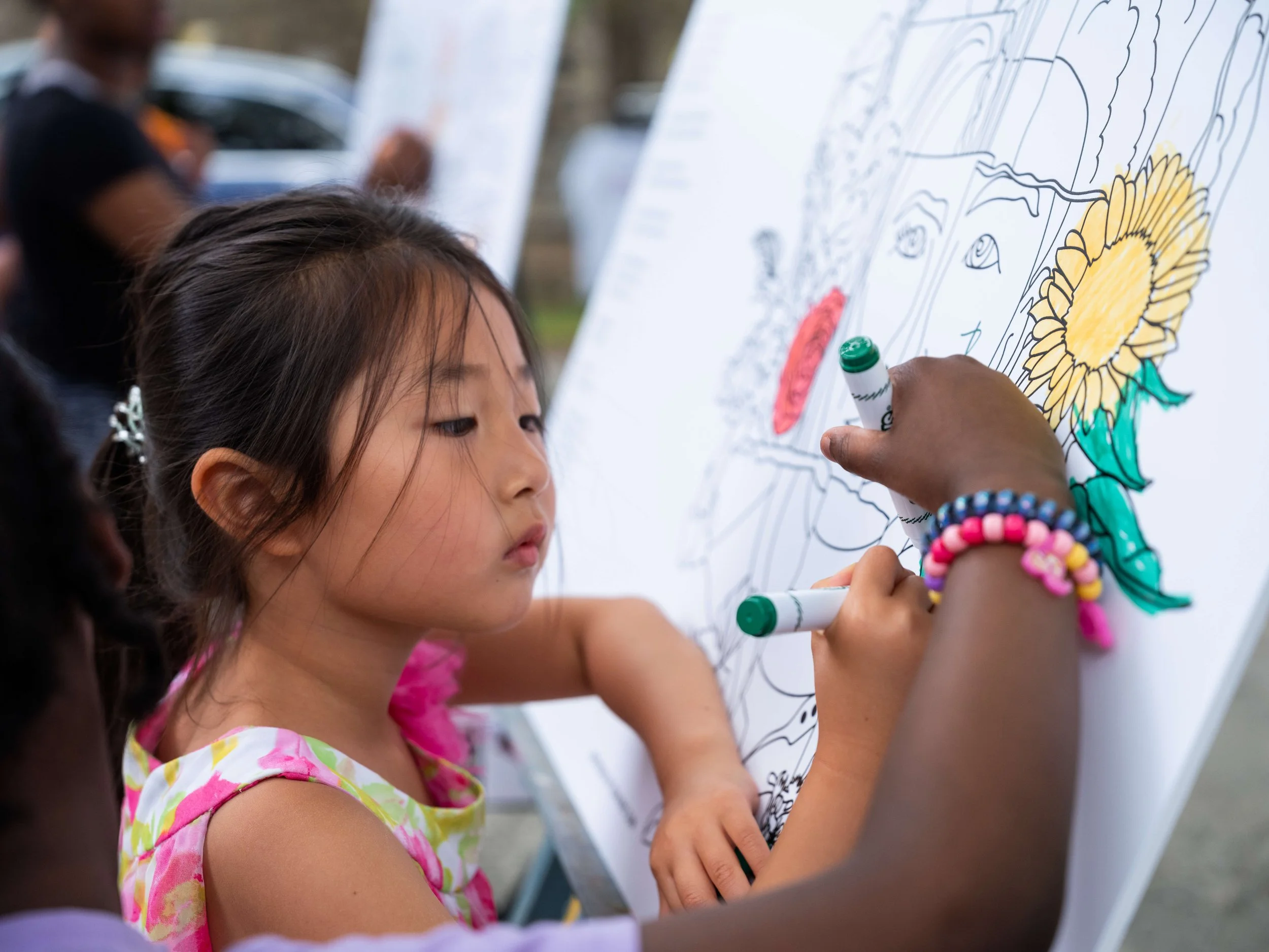 A young Asian and a young Black girl color together on a large collaborative art board featuring artwork from the ABC's of AAPIs Coloring Book.