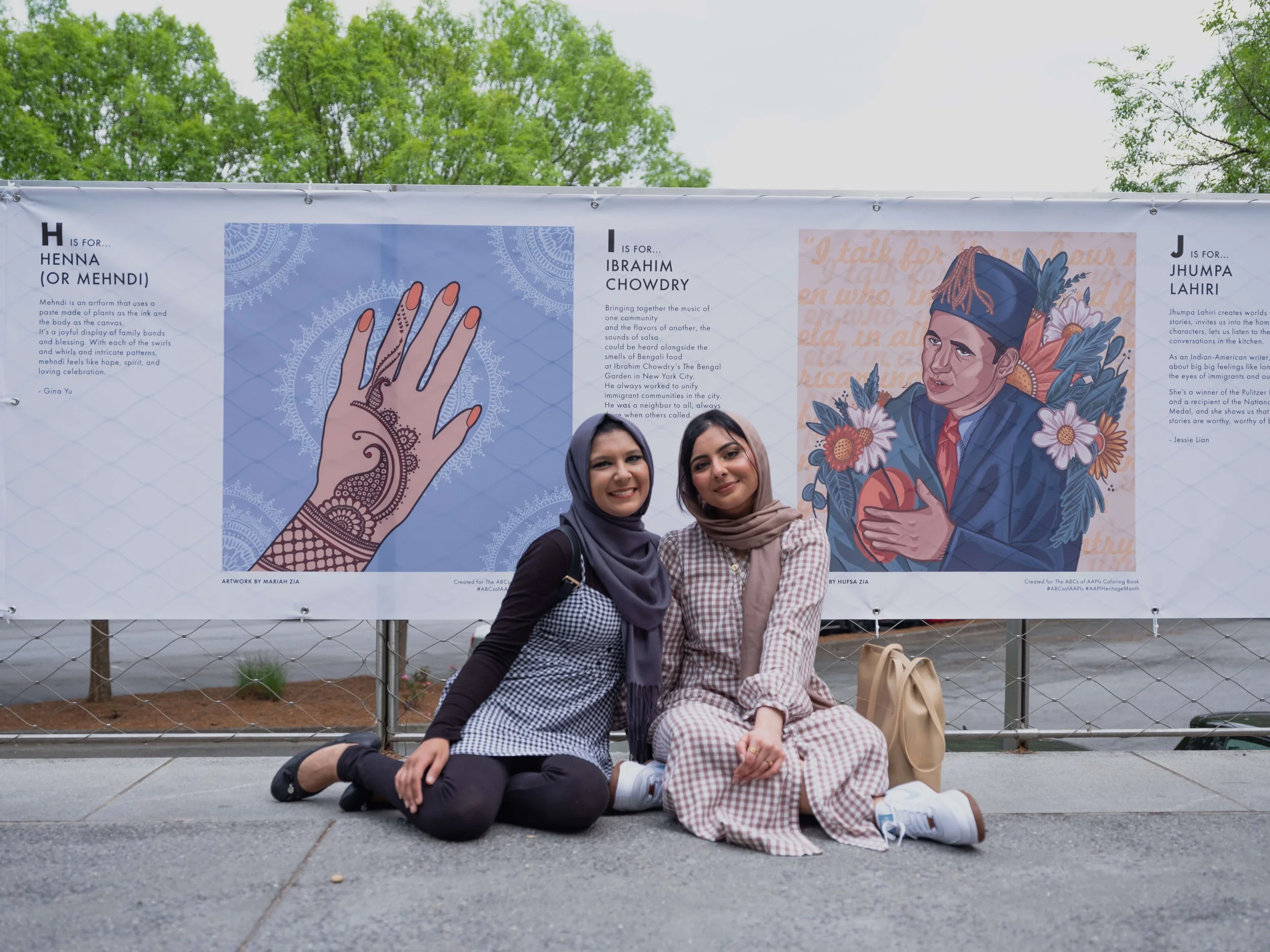 Artists Mariah Zia and Hufsa Zia sitting infront of the their artwork from the ABC's of AAPIs coloring book that is being displayed on the Atlanta Beltline behind them.