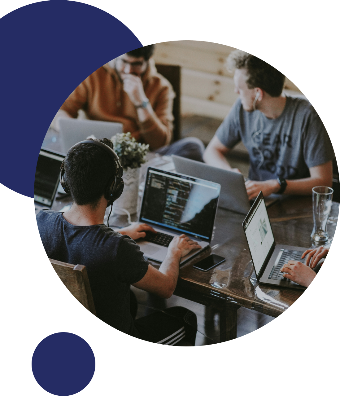 Group of young people working on laptops and computers at a wooden table, with one person wearing headphones.