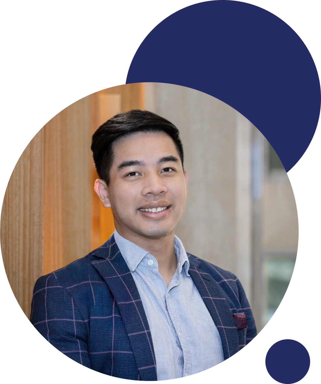 A young Asian man with short, dark hair smiling, wearing a blue plaid blazer over a light blue button-down shirt, standing indoors against a wooden wall with a window in the background.