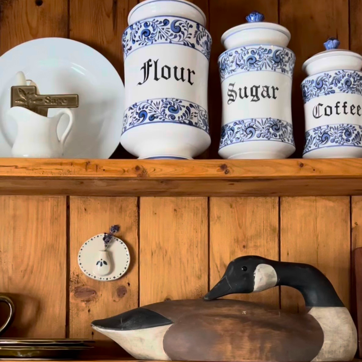 Kitchen shelf with vintage ceramic containers labeled Flour, Sugar, and Coffee, a small clock, a wall vase, and a wooden duck decoy. There is also a ceramic dish with a small pitcher labeled Sage, and a small decorative spoon hanging on the clock.