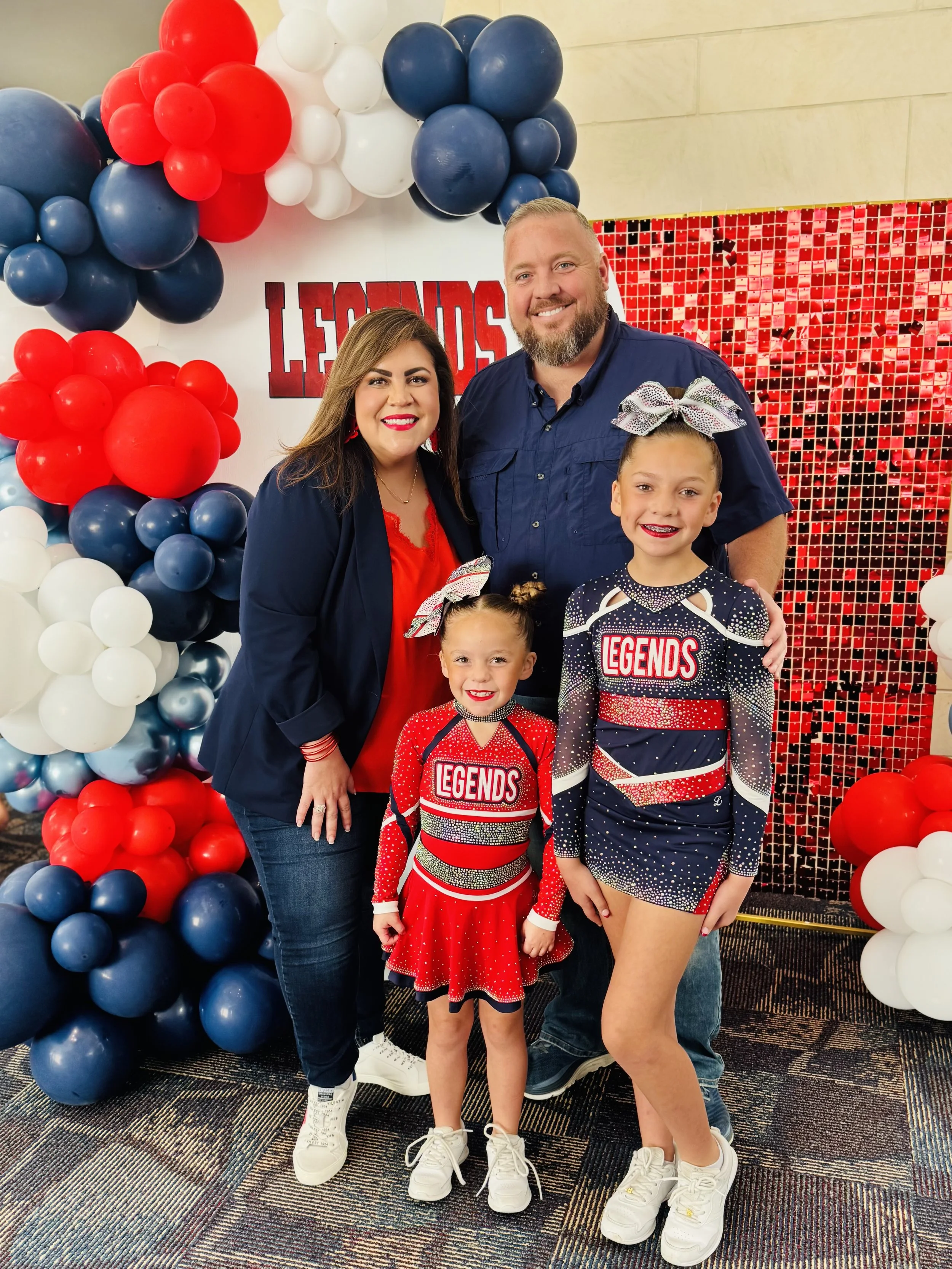 A family of four standing in front of a patriotic decoration with red, white, and blue balloons and a sign that says 'Legends'. The two daughters are dressed in cheerleading uniforms, and the parents are smiling nearby.