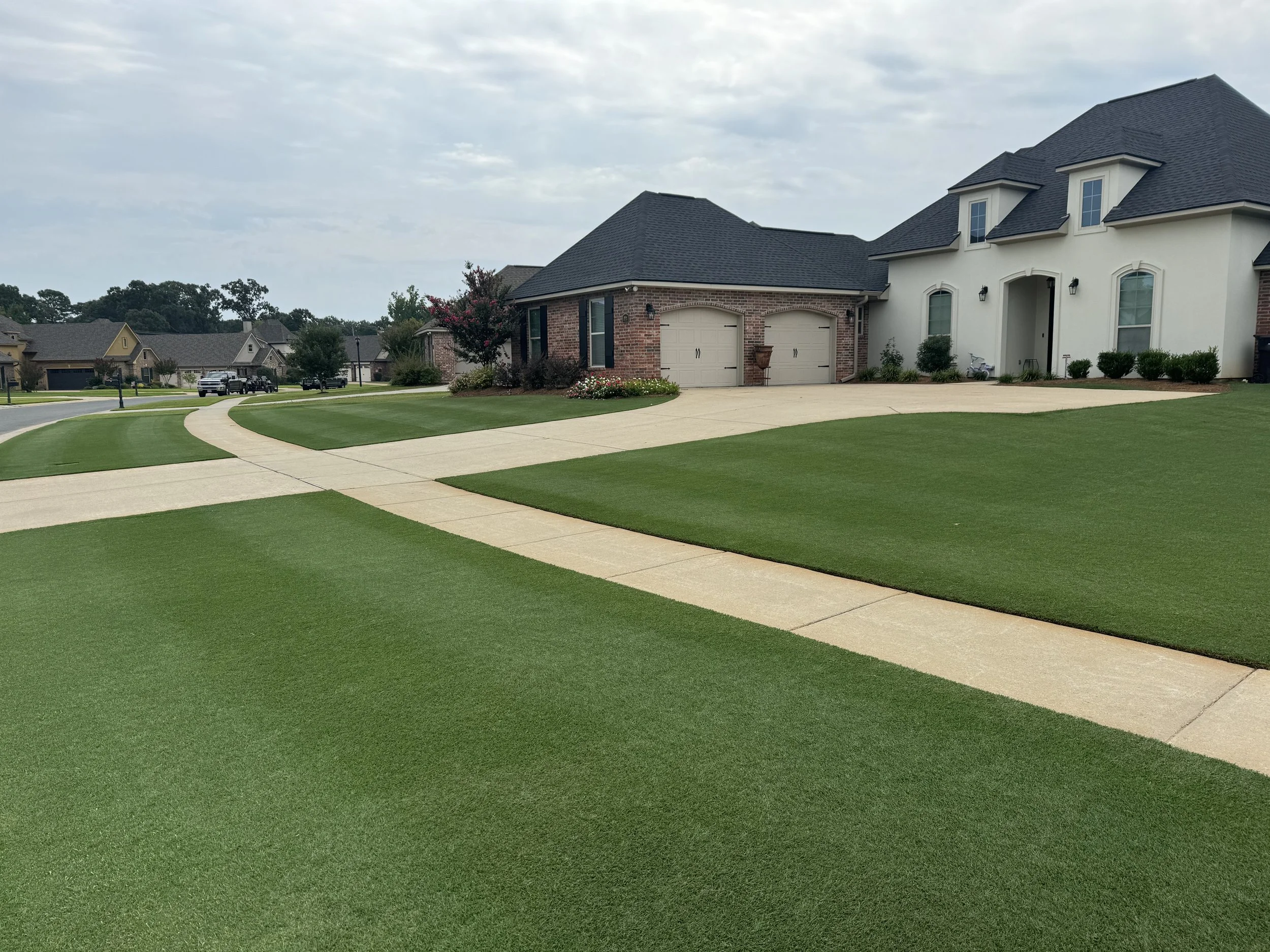 Residential neighborhood with manicured lawns, concrete walkways, and large house with brick and white exterior.