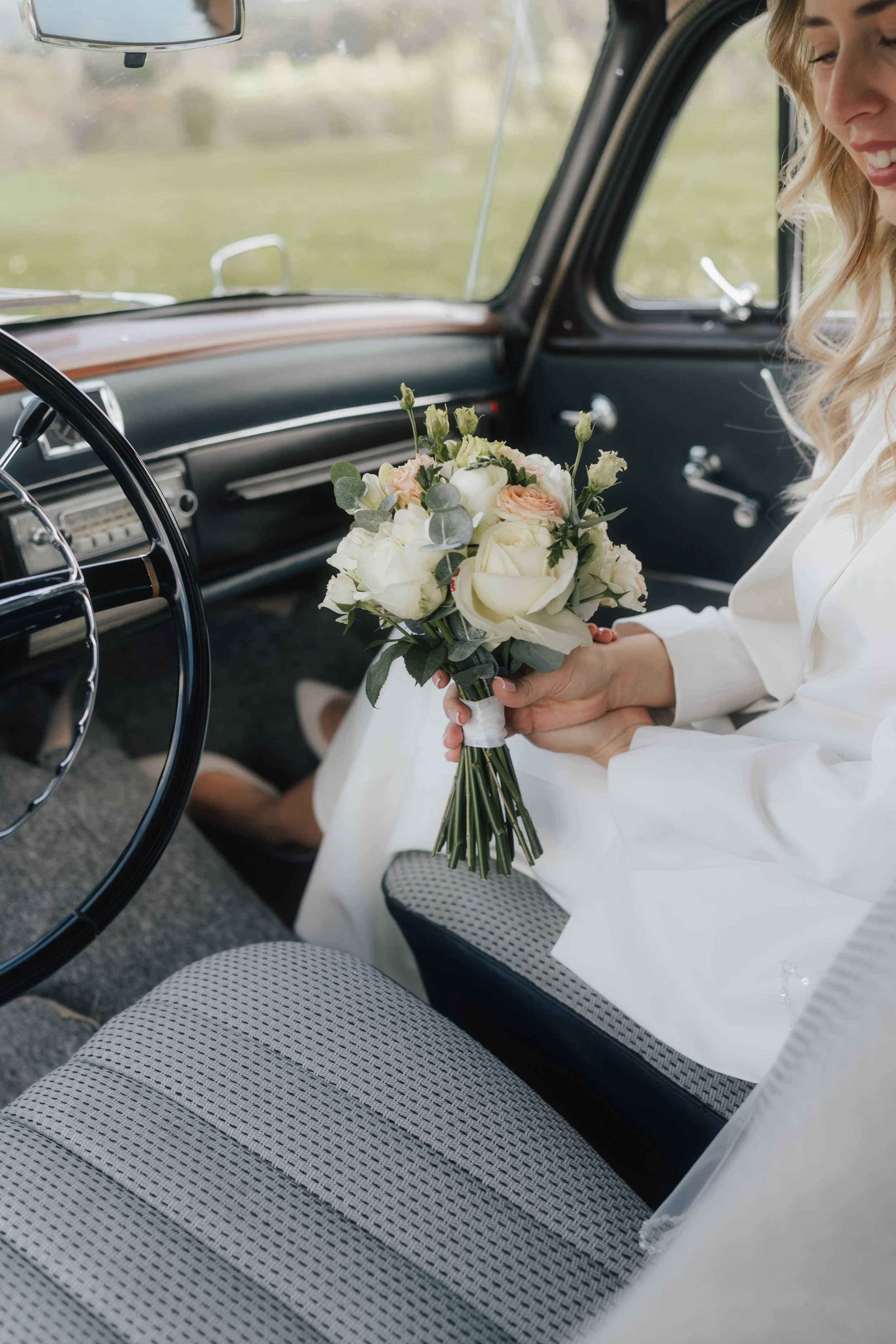 Eine Frau in einem weißen Kleid hält einen Blumenstrauß in einem Auto. Nur der obere Teil ihres Gesichts und ihre Hände sind sichtbar, sie lächelt.