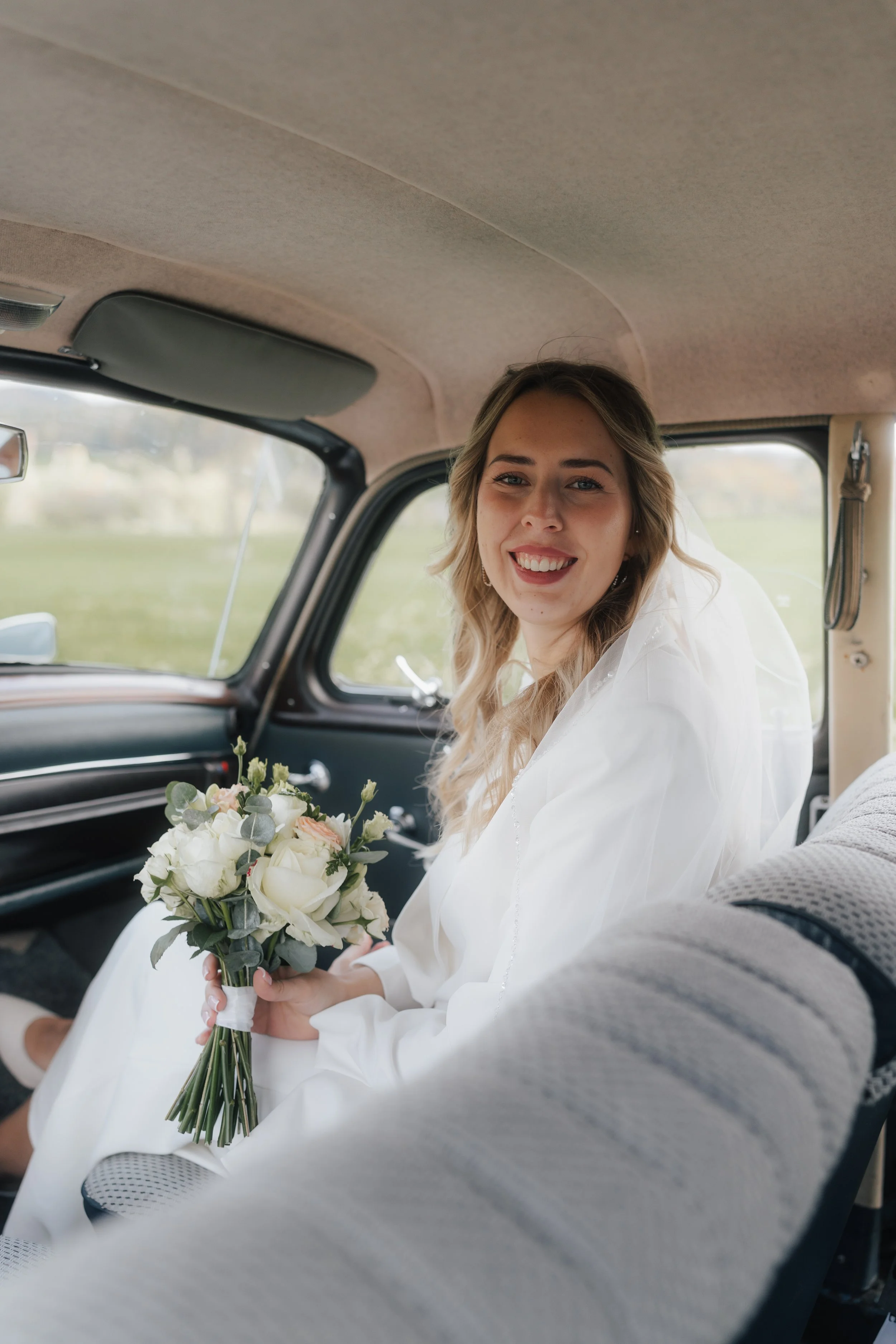 Junge Frau in Hochzeitskleid hält Brautstrauß im Auto und lächelt.