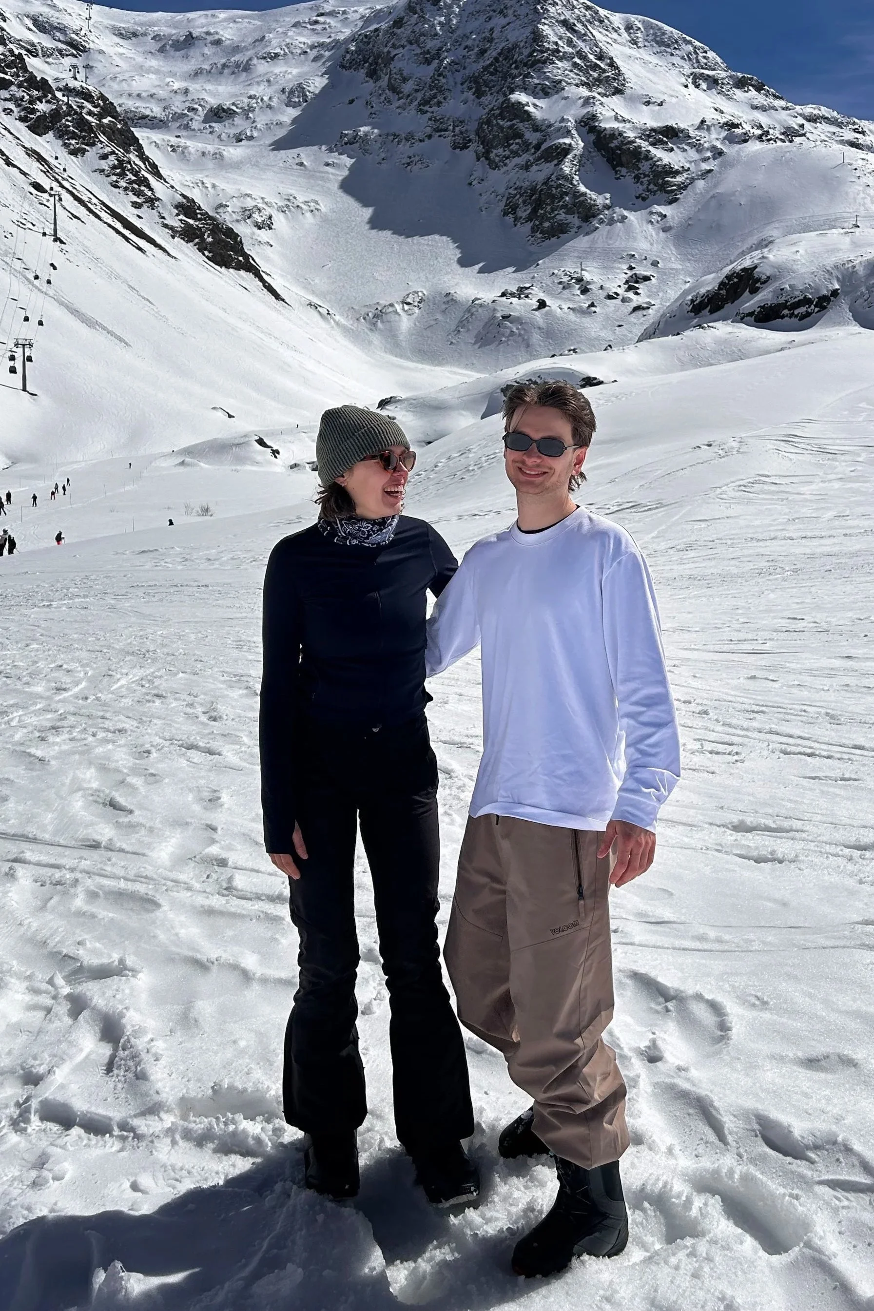 Zwei junge Menschen, eine Frau und ein Mann, lachen und posieren in einer verschneiten Berglandschaft mit Skiliften im Hintergrund.