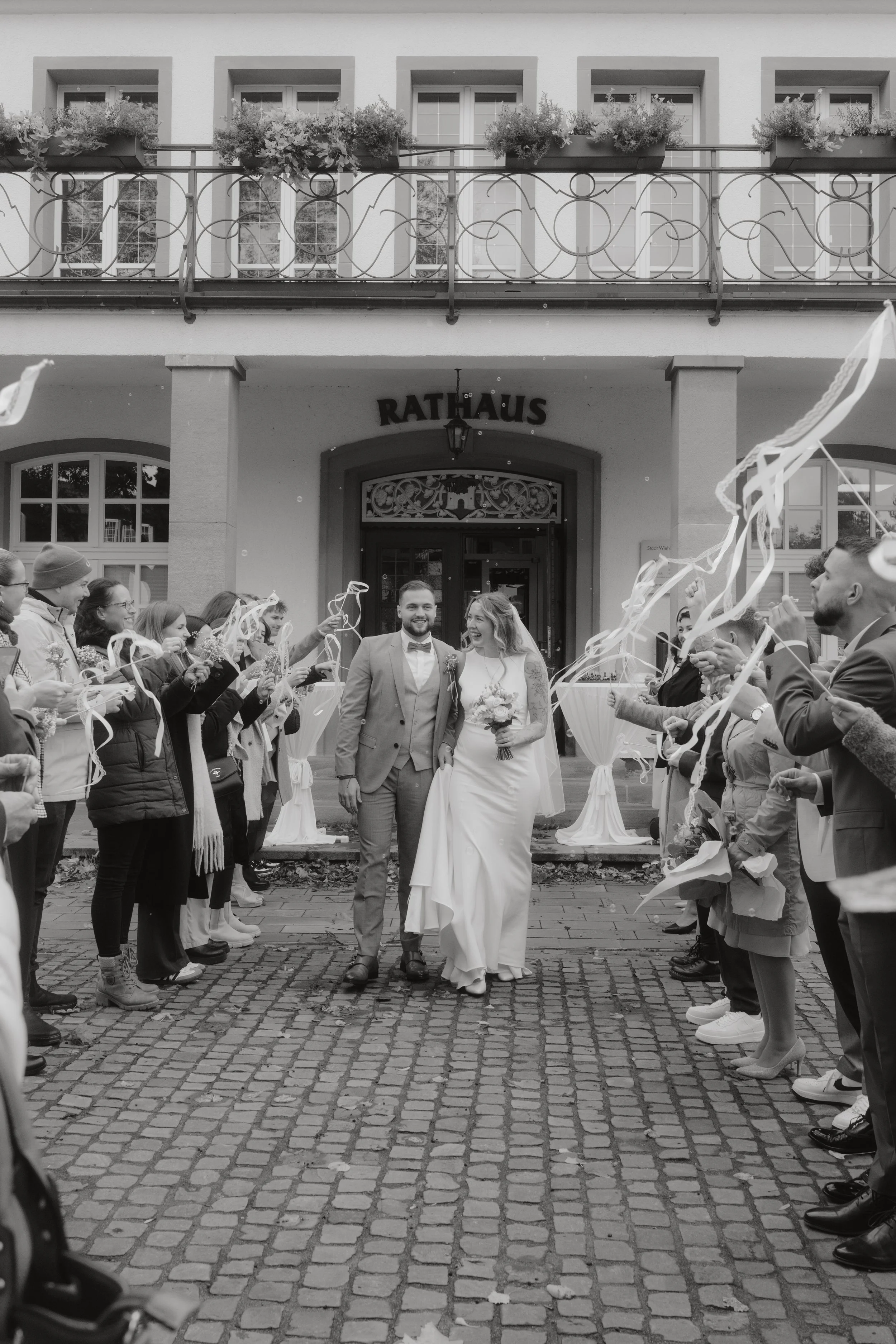 Ein Brautpaar verlässt eine Zeremonie, umgeben von feiernden Gästen, vor einem Gebäude mit der Aufschrift 'Rathaus'.