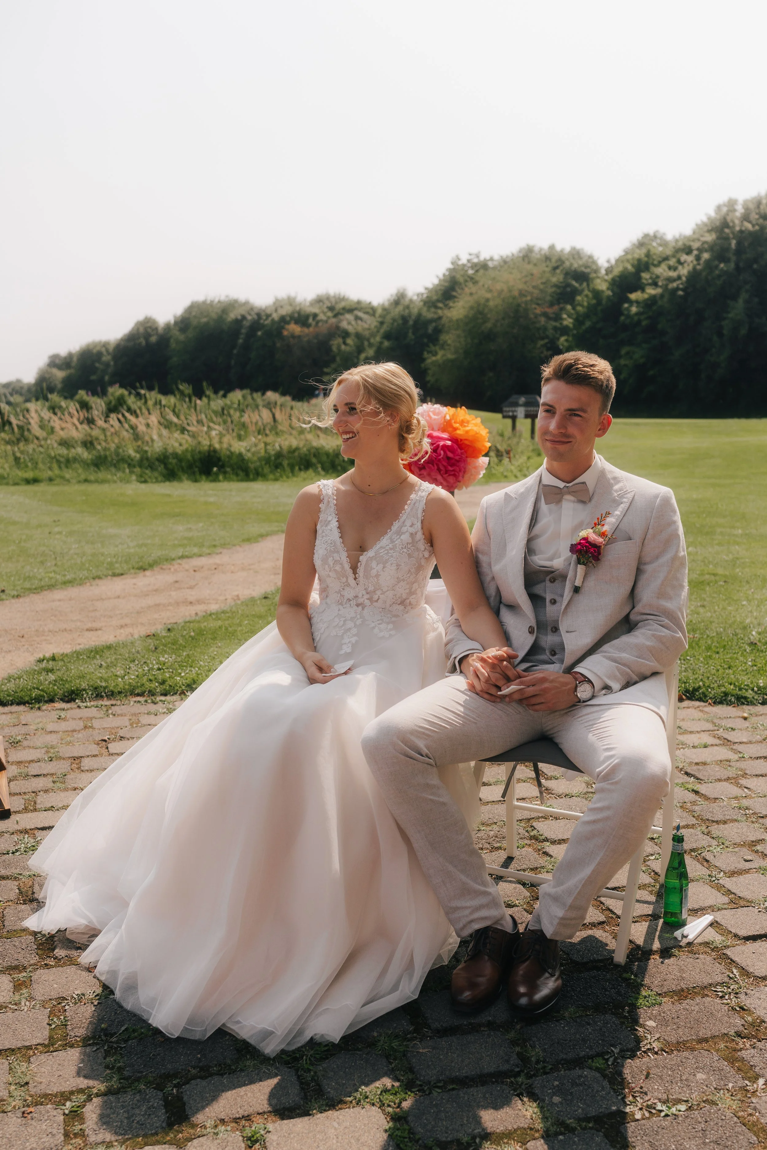 Ein frisch vermähltes Paar sitzt im Freien bei ihrer Hochzeit, die Braut trägt ein weißes Hochzeitskleid mit Spitze, der Bräutigam einen hellen Anzug mit Fliege, im Hintergrund sind bunte Blumen und grüne Bäume zu sehen.