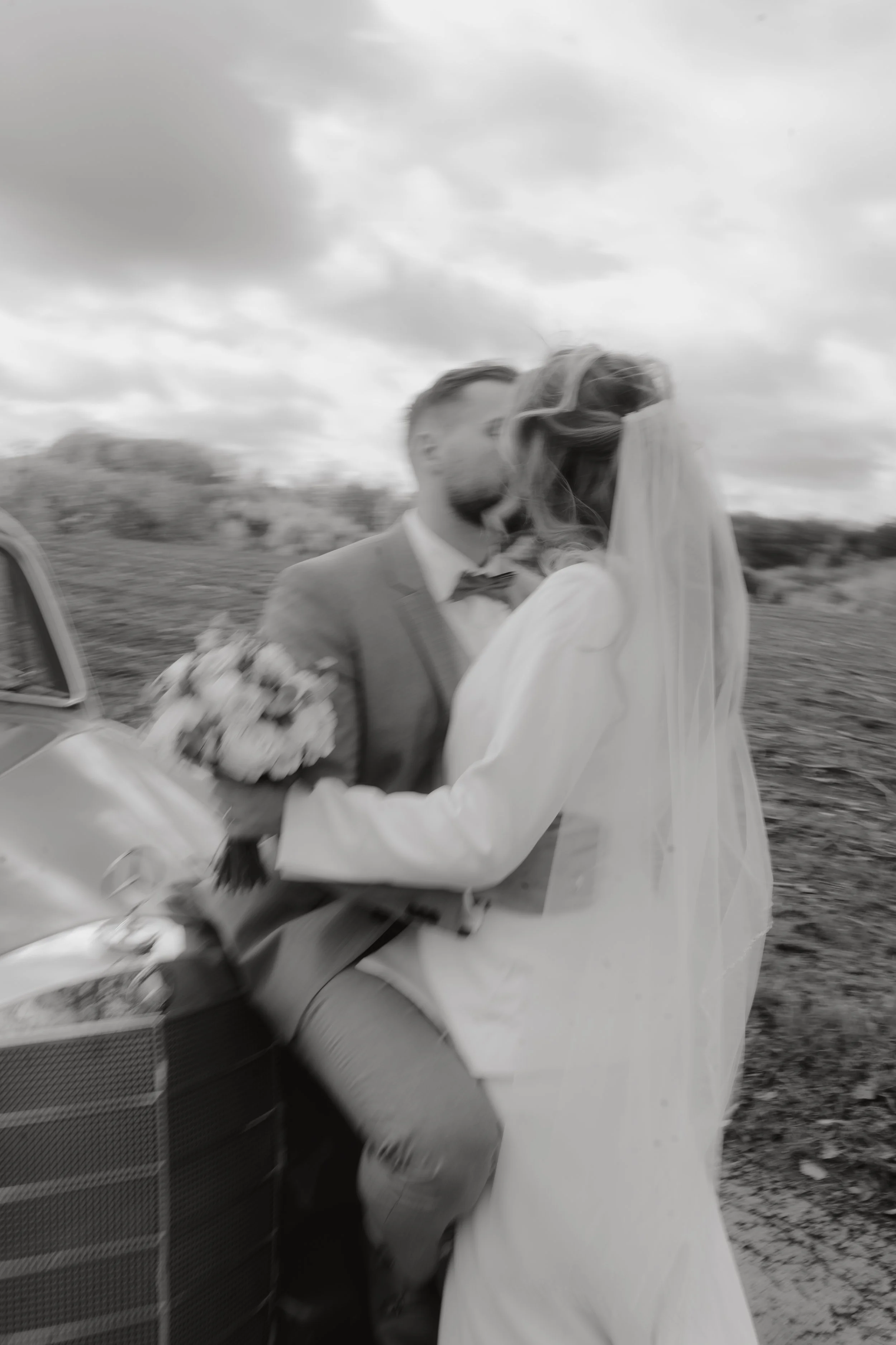 Ein sich küssendes Brautpaar in Schwarz-Weiß-Fotografie, das an einem Auto im Freien steht, mit bewölktem Himmel im Hintergrund.