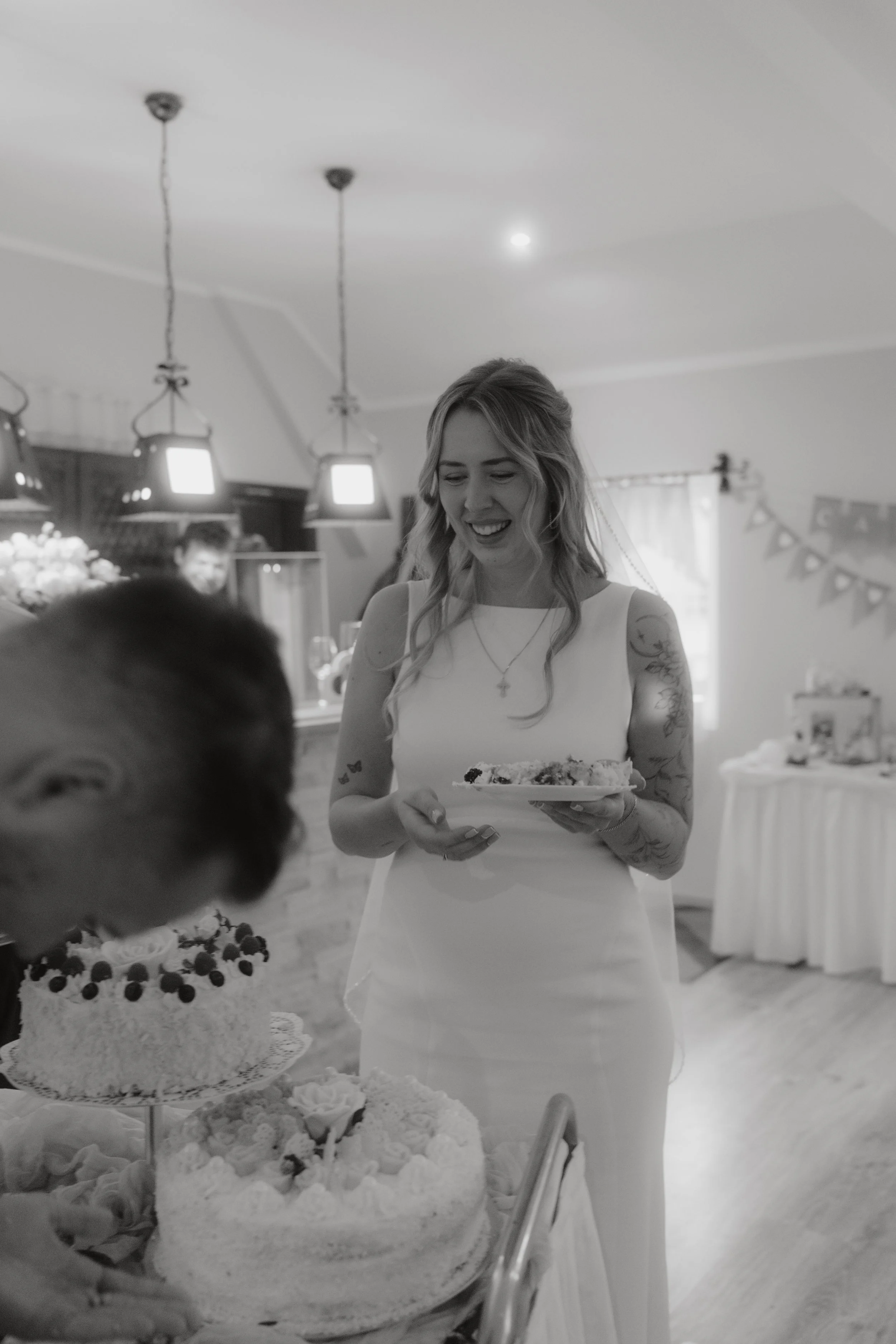 Eine junge Frau in einem weißen Kleid hält ein Stück Kuchen in der Hand bei einer Feier, im Vordergrund ist ein großer, reich verzierter Geburtstagkuchen mit Früchten.