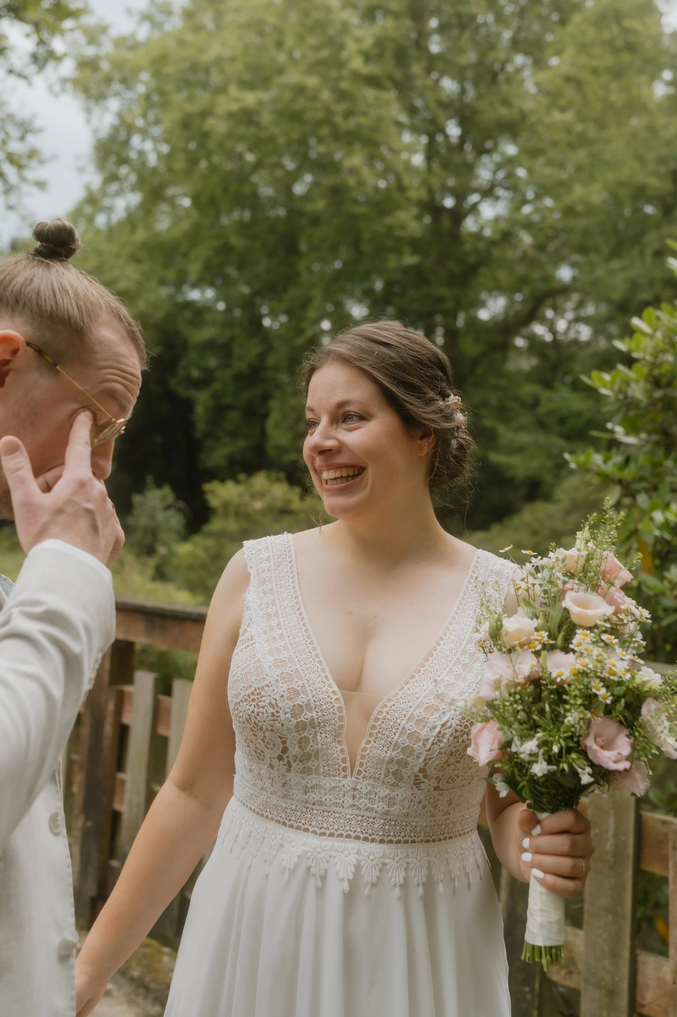 Eine lachende Braut mit Brautstrauß, im Gespräch mit einem Mann, an einem grünen, bewaldeten Ort.