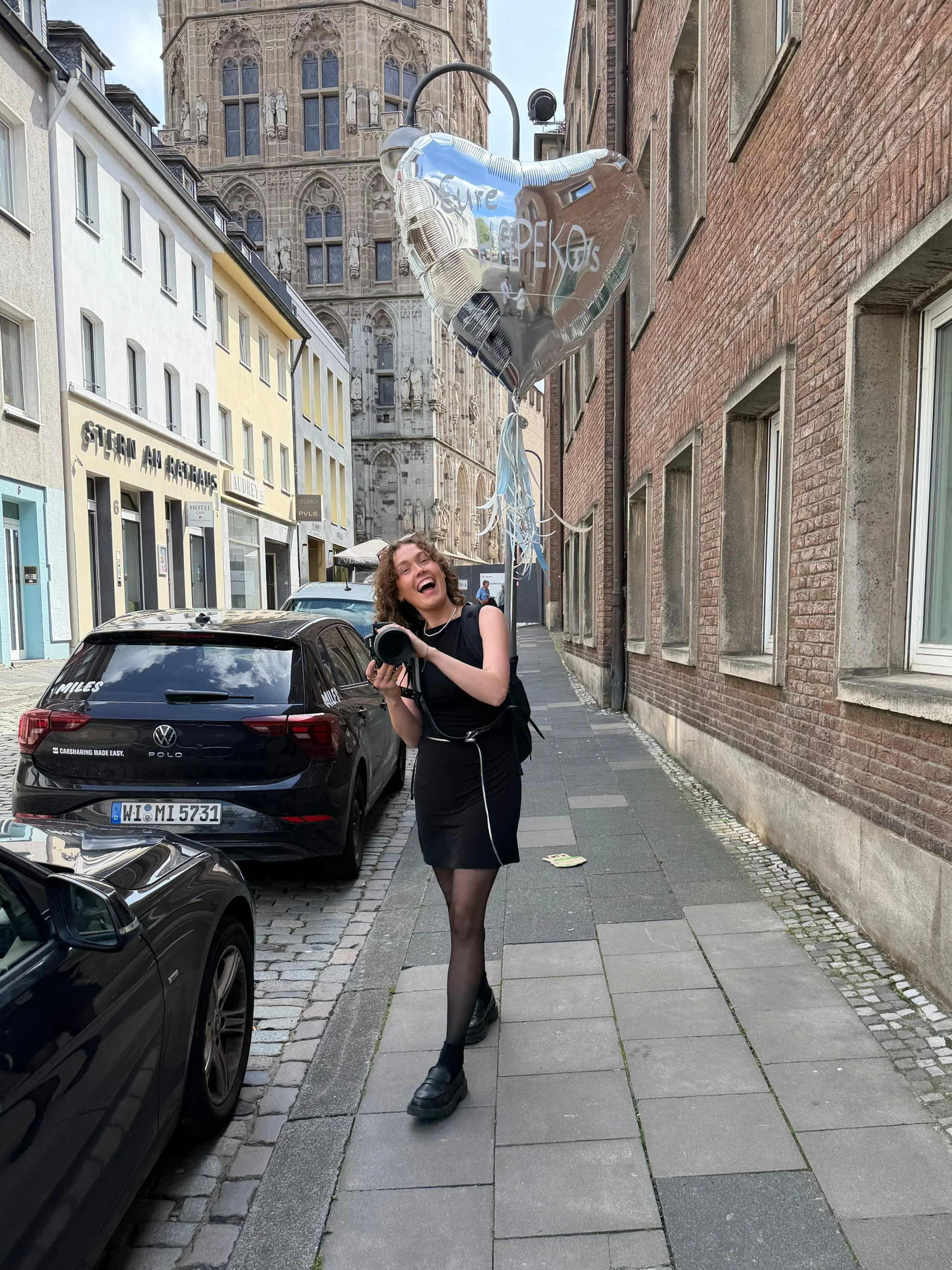 Eine lachende Frau mit dunkler Kleidung, schwarzen hohen Schuhen und einem schwarzen Rucksack läuft auf einem Bürgersteig neben geparkten Autos, während sie eine Herzballon mit der Beschriftung 'Eure Hochzeits' hält, vor einer historischen Kirche in 