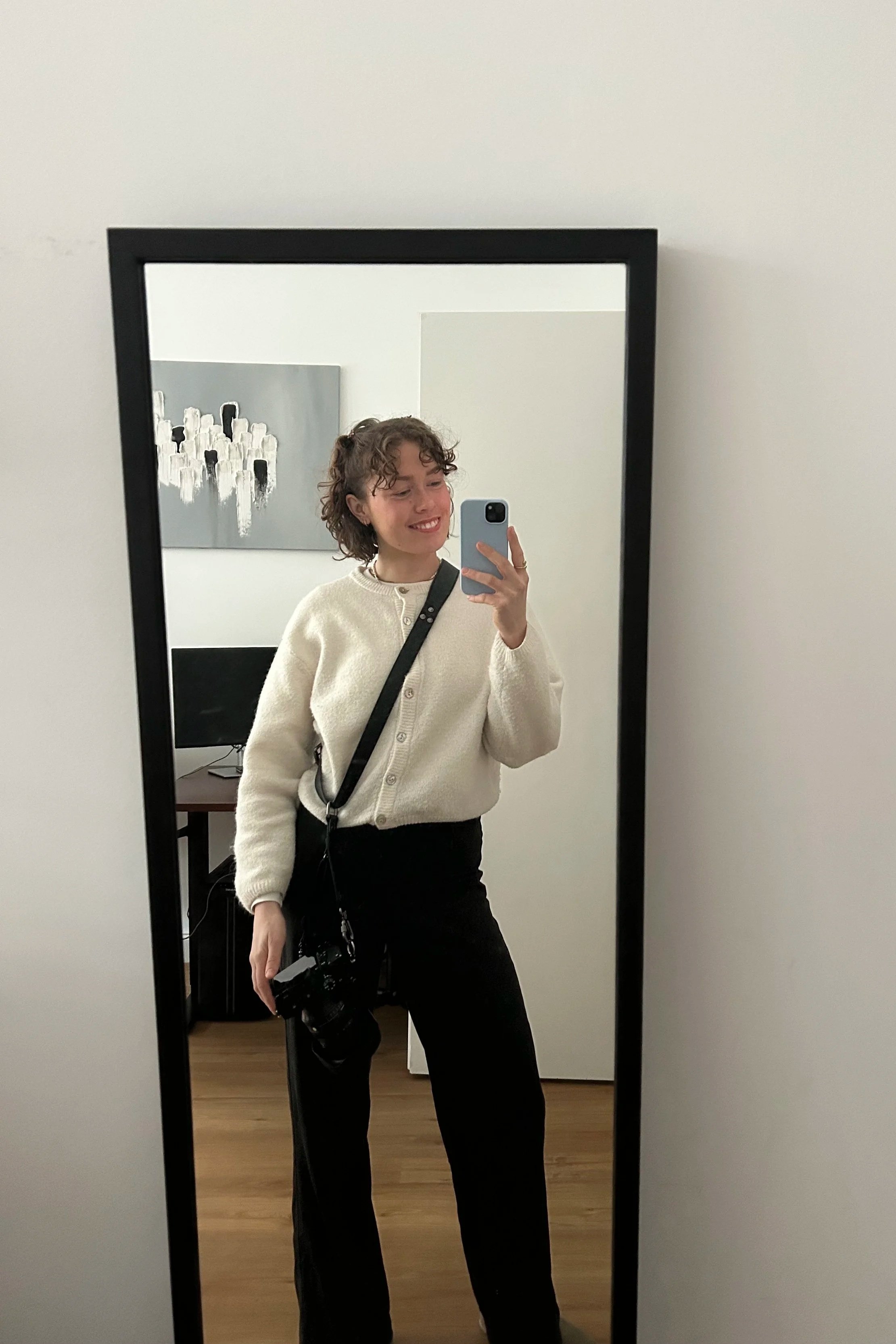 Eine junge Frau macht ein Selfie in einem Spiegel. Sie trägt eine weiße Strickjacke, schwarze Hosen und hat einen schwarzen Rucksack. Im Hintergrund ist ein Fernseher und ein abstraktes Kunstwerk an der Wand.