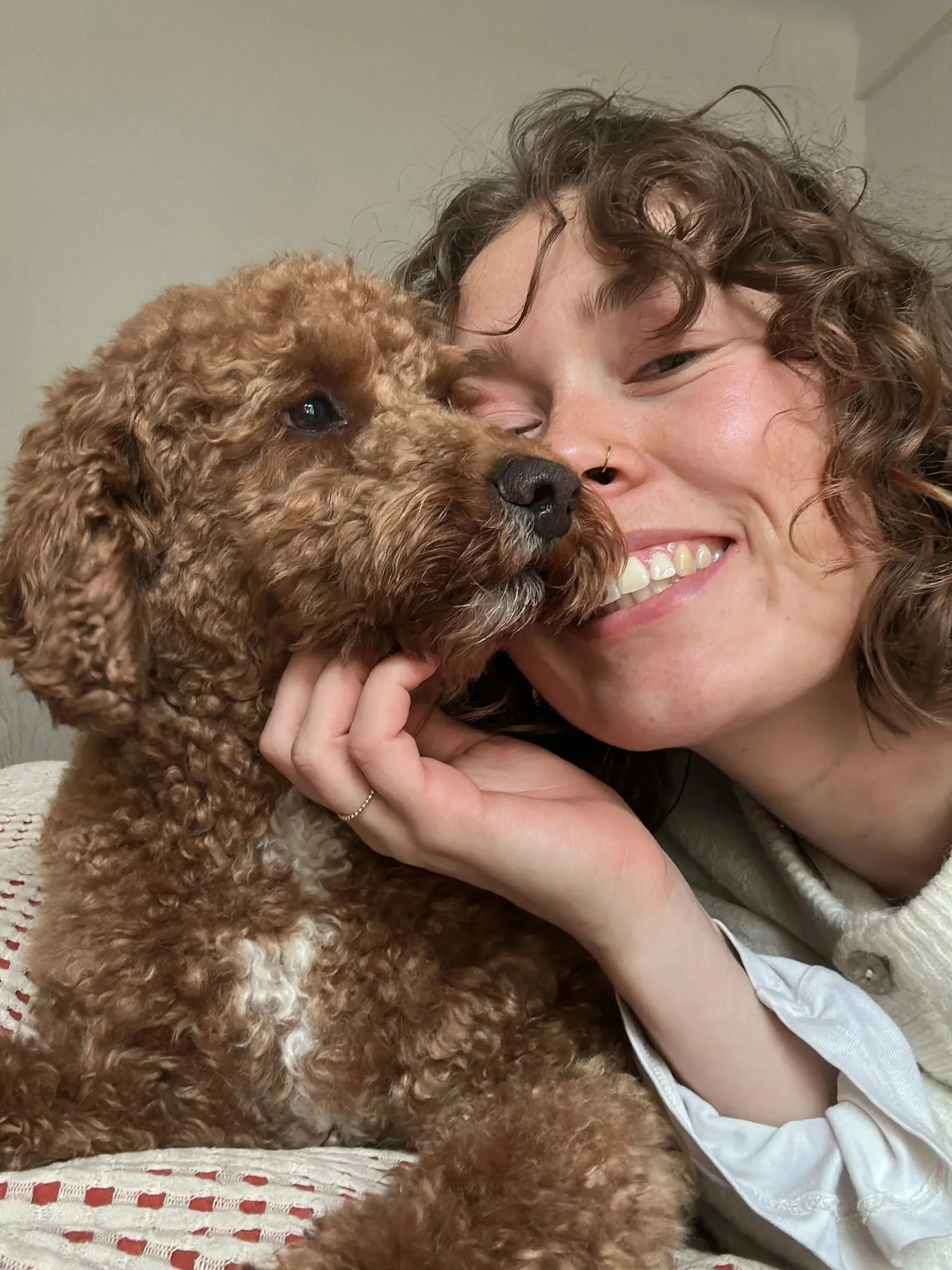 Ein lachender Mensch mit Locken und Piercing im Nasenbereich, der einen braunen Lockenhund streichelt, sowohl die Person als auch der Hund kuscheln eng miteinander.