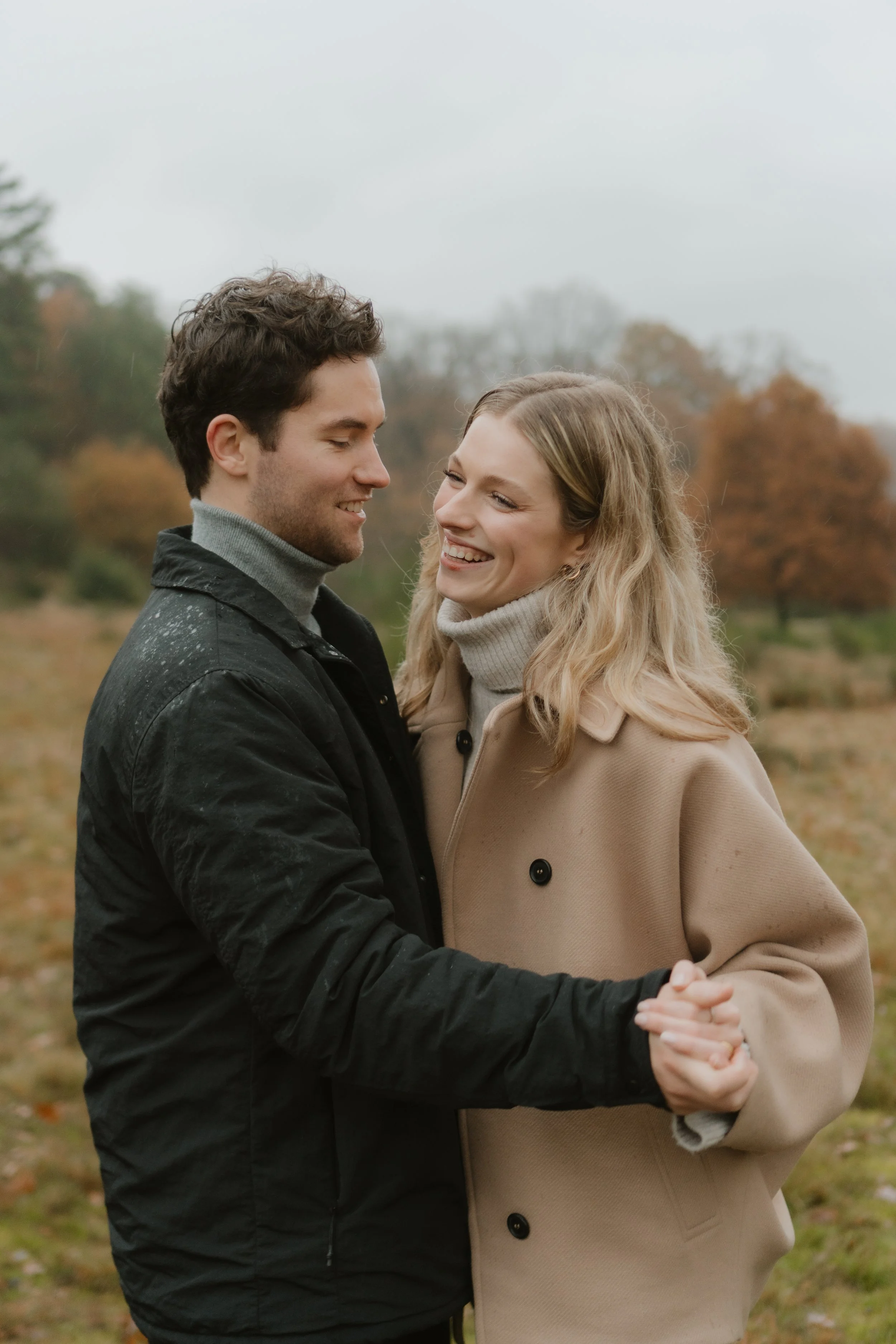 Ein Paar lächelt und umarmt sich draußen im Herbst. Im Hintergrund Bäume mit buntem Laub.