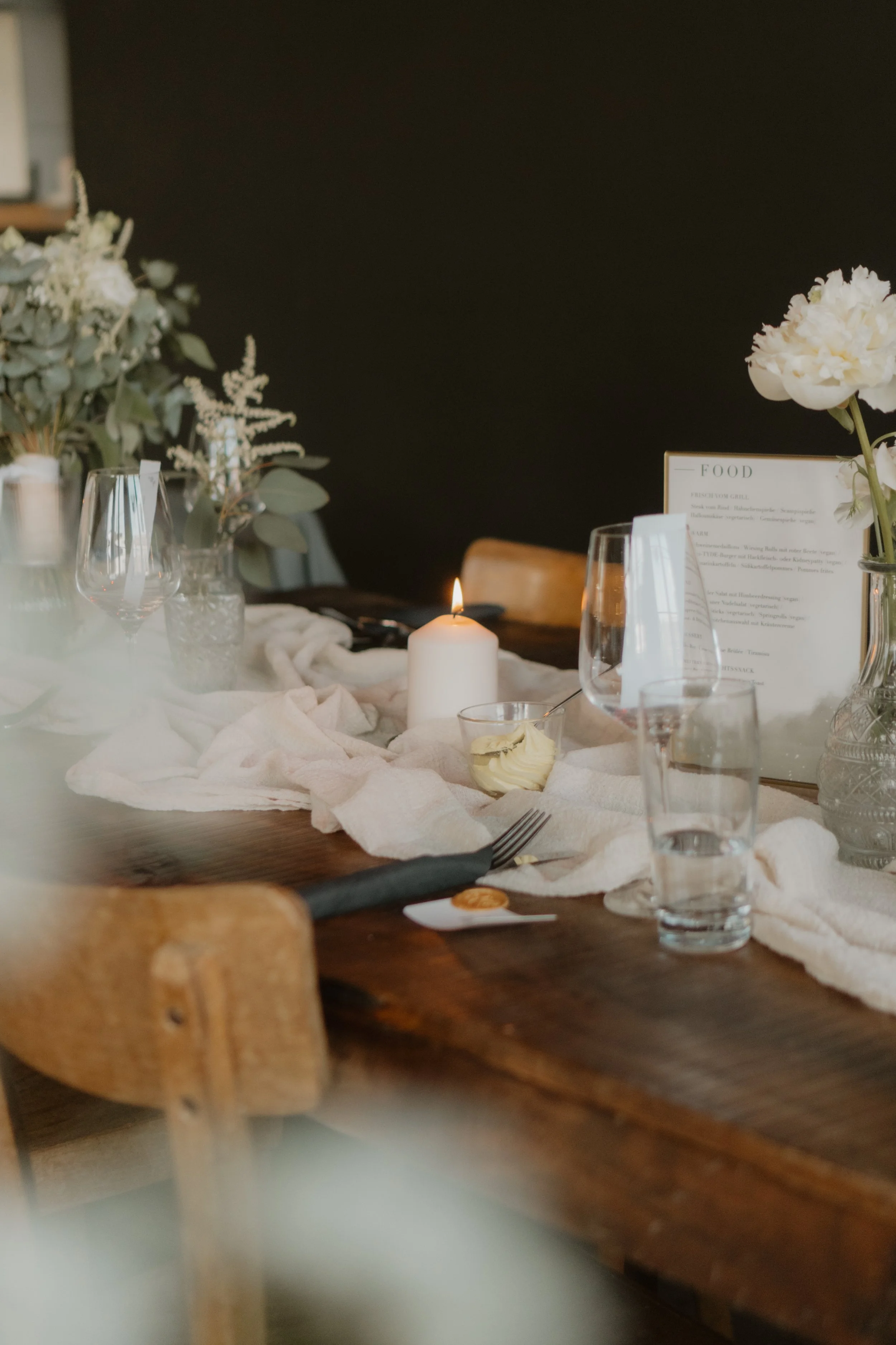 Ein gedeckter Esstisch mit weißen Stoffläufern, Kerzen, Wein- und Wassergläsern, Blumen in Vasen, einem Menü und Besteck, in einer gemütlichen Umgebung.