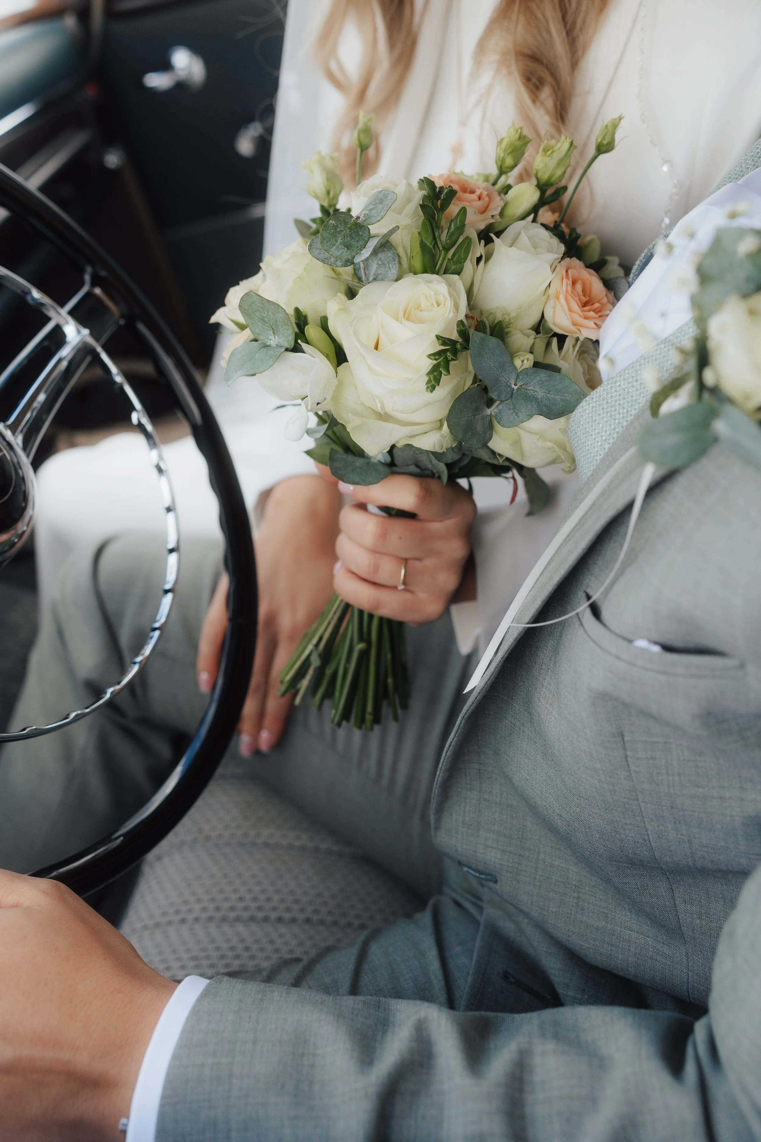 Person hält einen Brautstrauß aus weißen und peachfarbenen Rosen mit Eukalyptus in der Hand, während sie in einem Auto sitzt, vermutlich an ihrem Hochzeitstag.