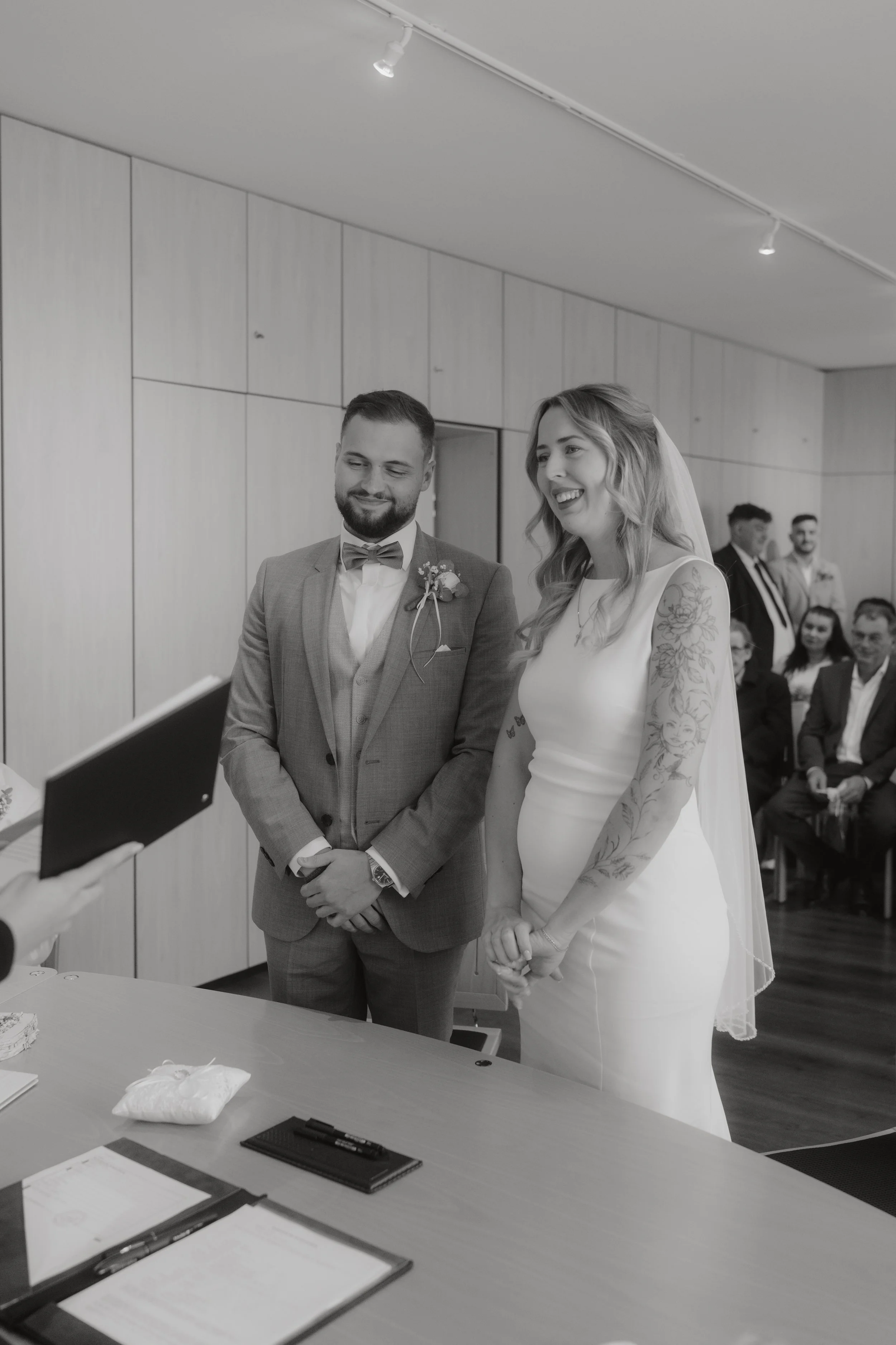 Braut und Bräutigam bei ihrer Hochzeit, in einem Raum mit mehreren Gästen, im Moment der Trauung, im schwarzen und weißen Foto.