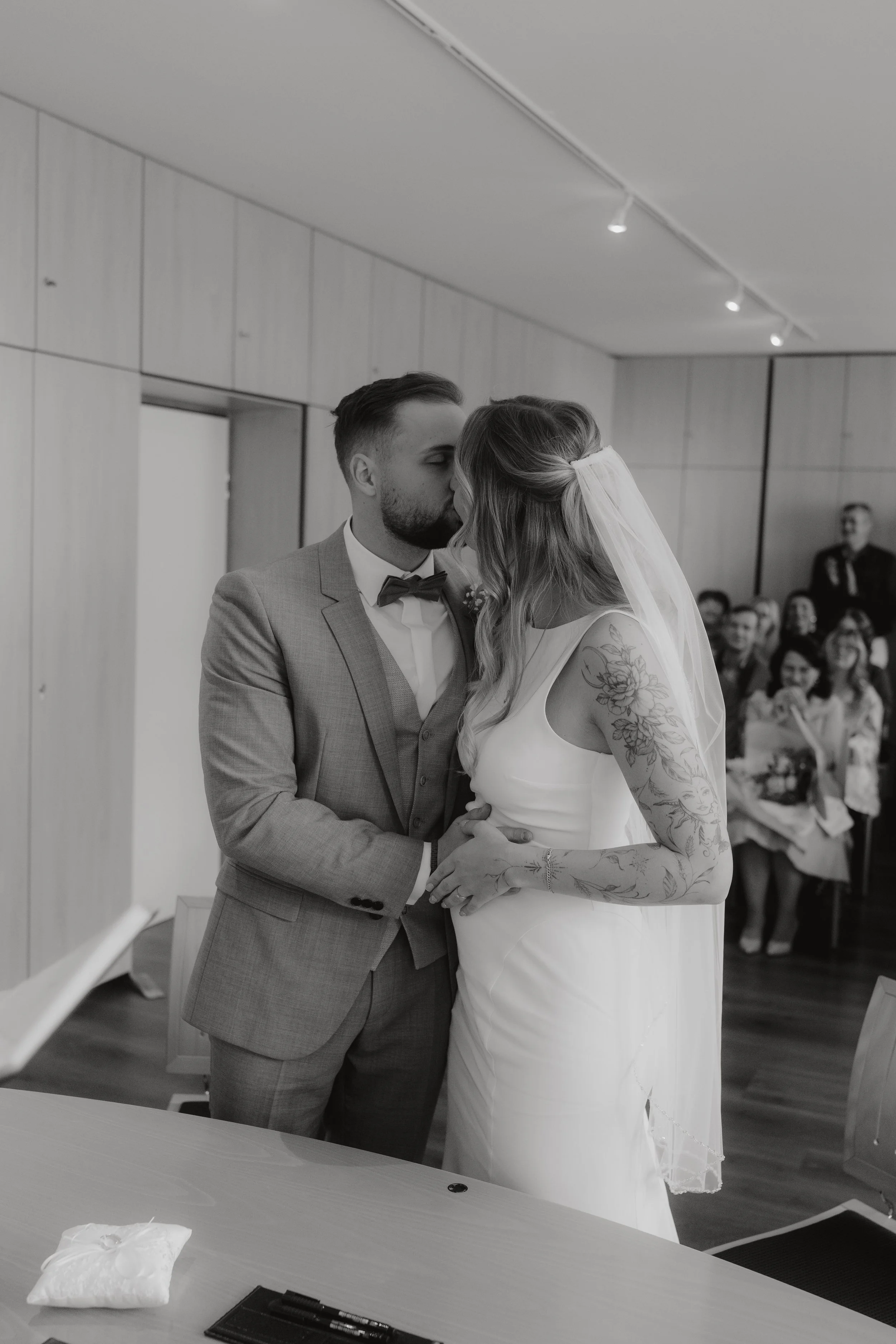 Ein Brautpaar bei ihrer Hochzeit, während sie sich küssen, mit Gästen im Hintergrund, in einem Raum mit Holzverkleidung.
