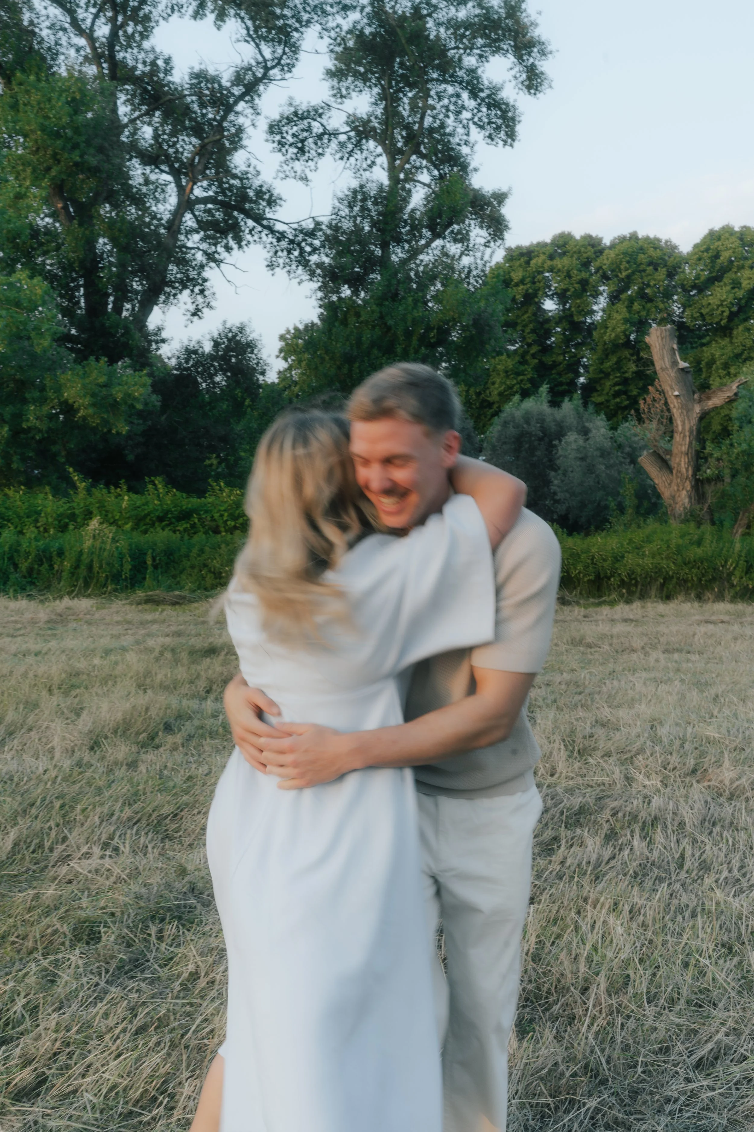 Paar umarmt sich lachend im Freien auf einer Wiese mit Bäumen im Hintergrund.