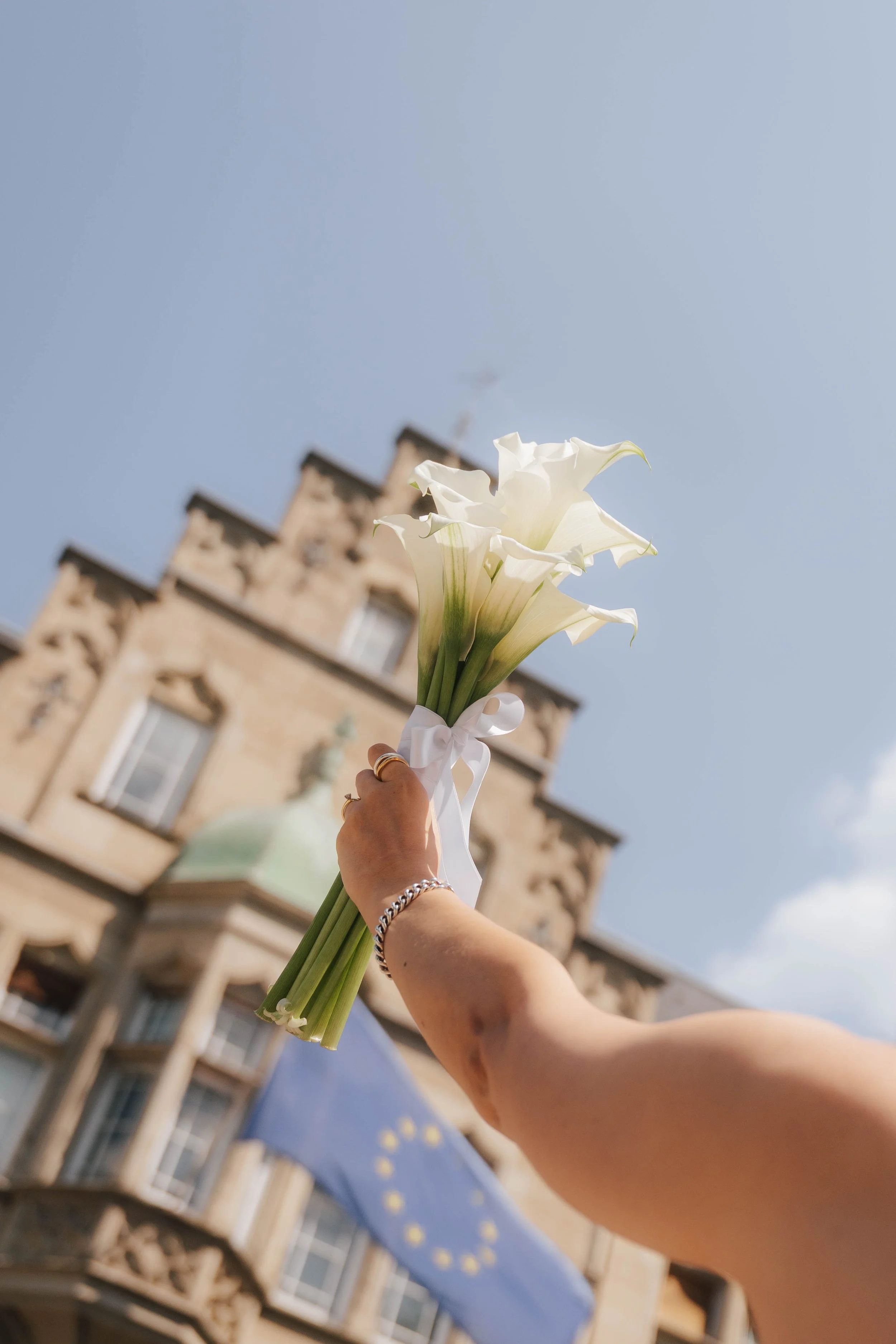 Person hält einen Strauß weißer Calla-Lilien vor einem alten Gebäude mit Turm im Hintergrund. Der Himmel ist blau und es ist sonnig.
