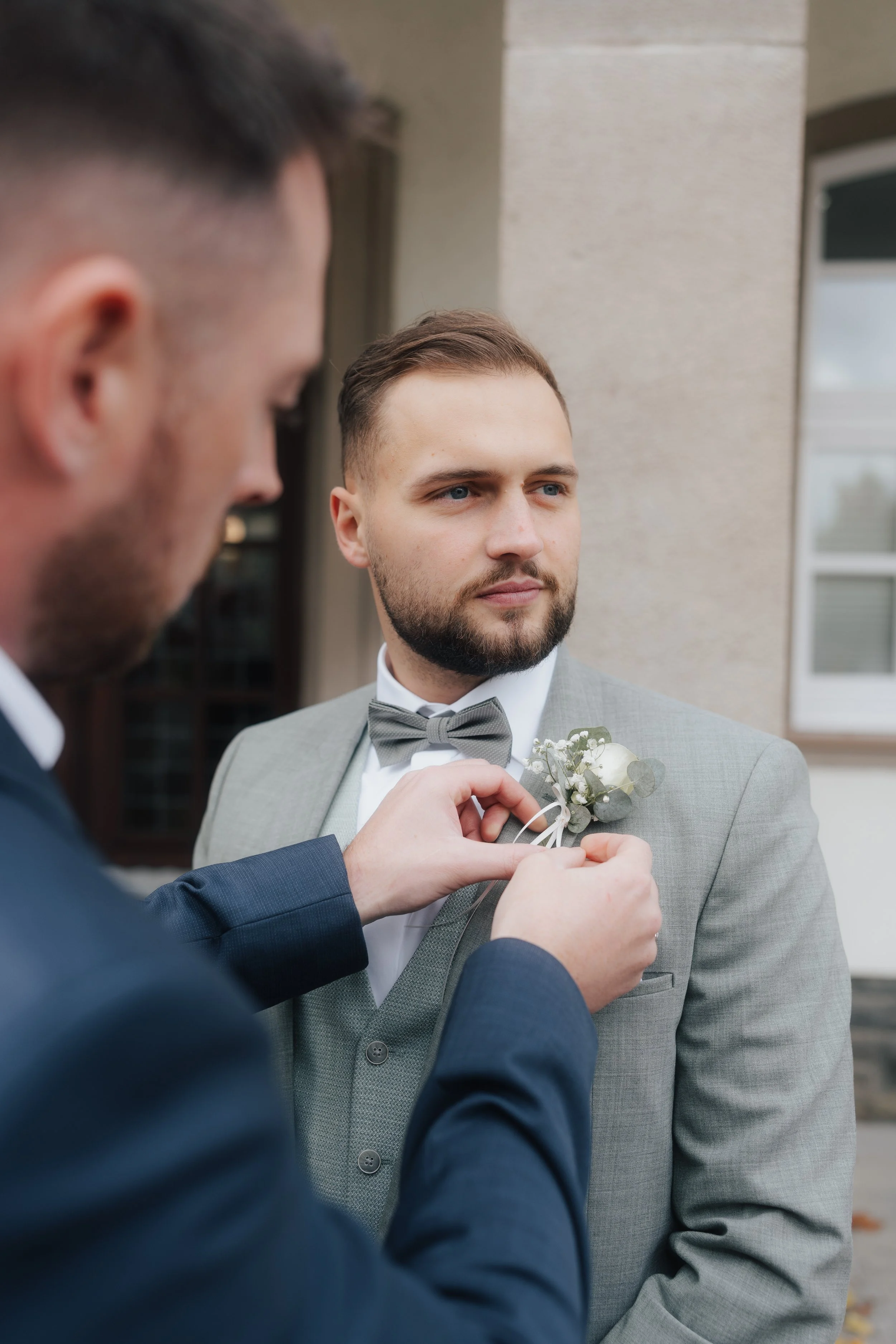 Ein Mann in einem grauen Anzug und Fliege, der an seinem Revers eine weiße Blumenkombination erhält, steht vor einem Haus.