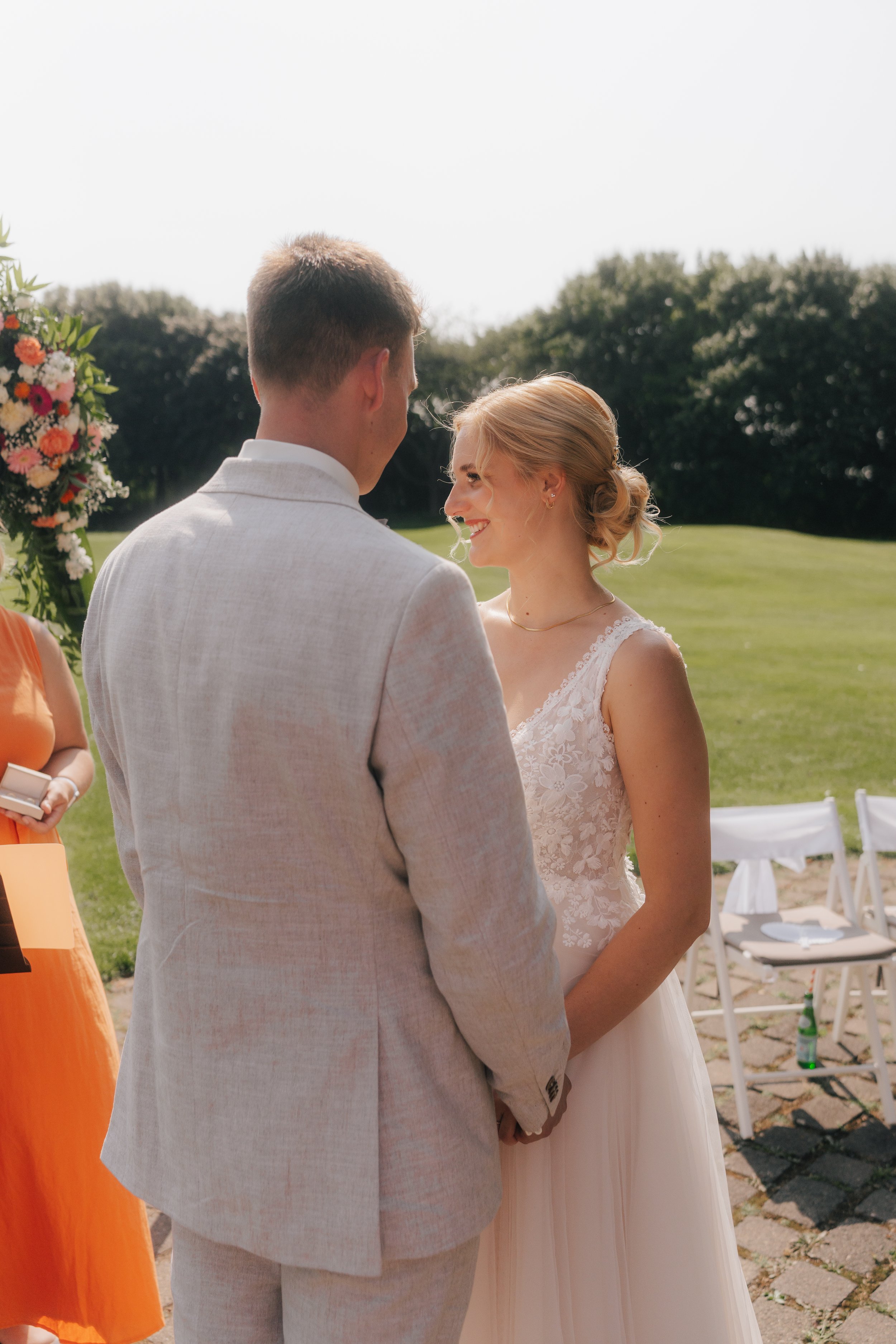 Ein Brautpaar bei einer Hochzeit im Freien, hält Händchen, die Frau trägt ein weißes Hochzeitskleid, der Mann einen hellen Anzug, im Hintergrund grüne Bäume und Stühle.