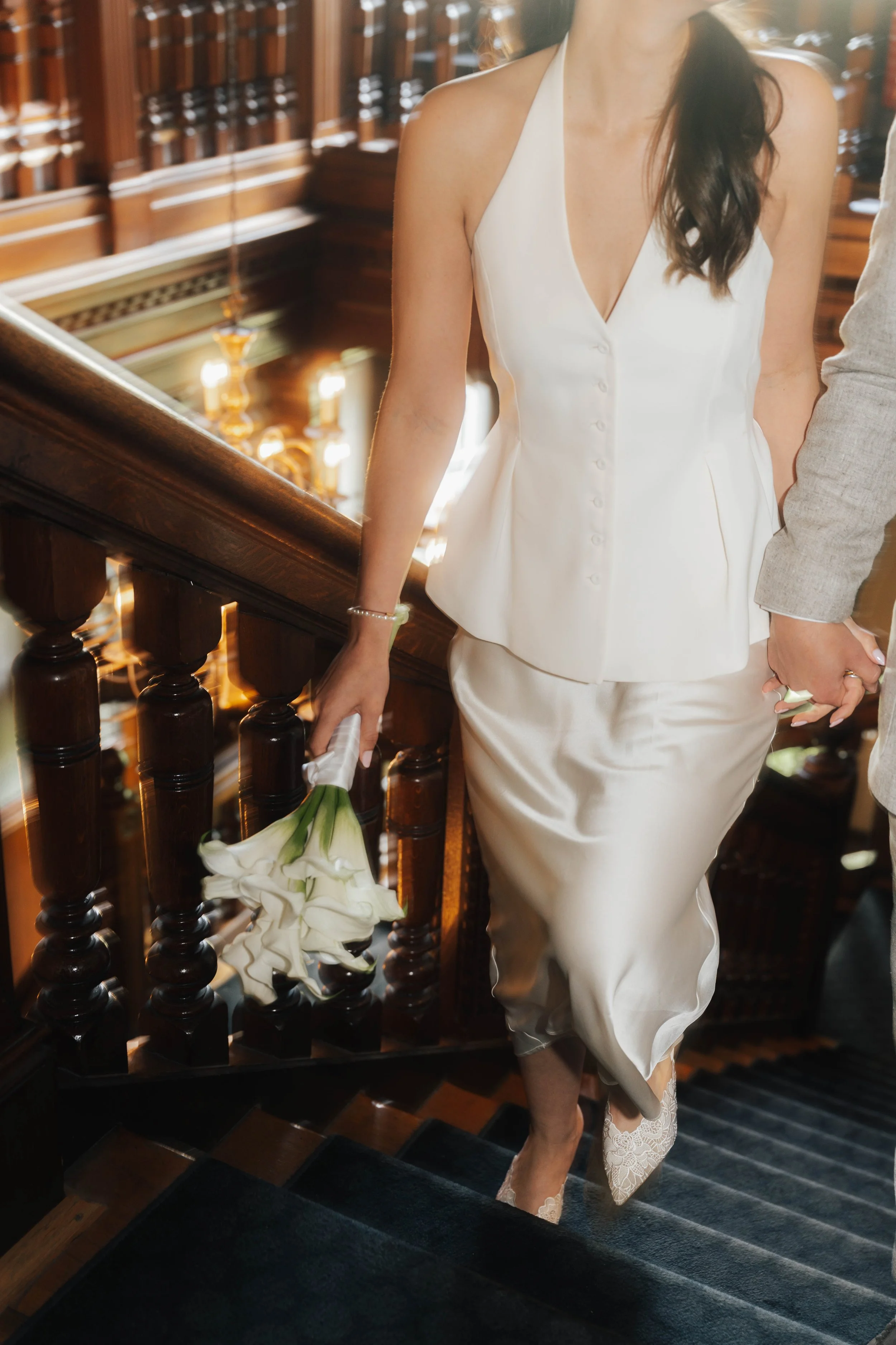Eine Frau in einem weißen Kleid steht auf einer dunklen Treppe, hält eine Bouquet aus weißen Blumen, umgeben von Holzgeländern und einer dekorativen, alten Innenarchitektur.